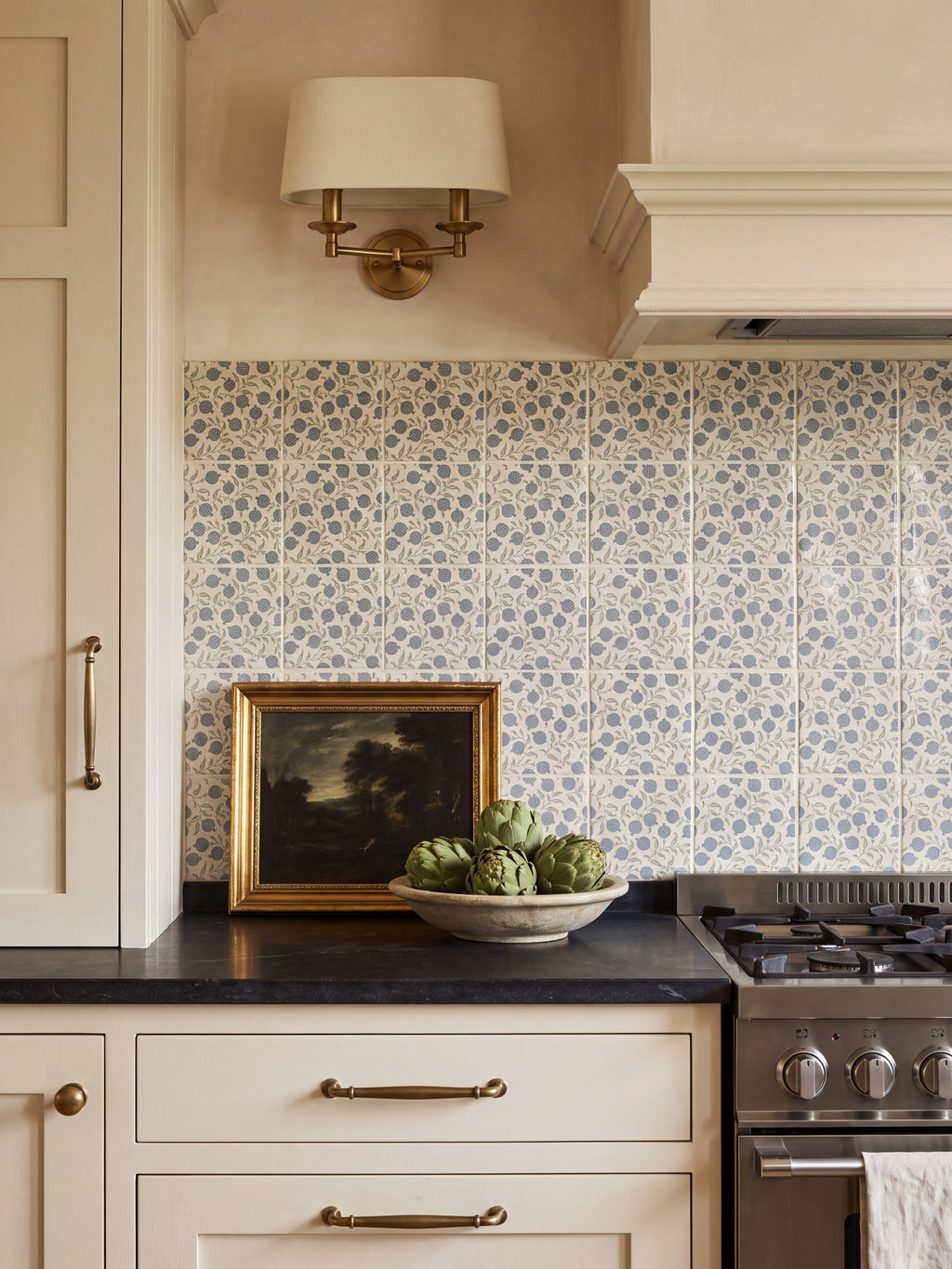 Anaar Pottery Blue tile splashback in a cream kitchen with black worktop, brass sconce and gilt-framed painting