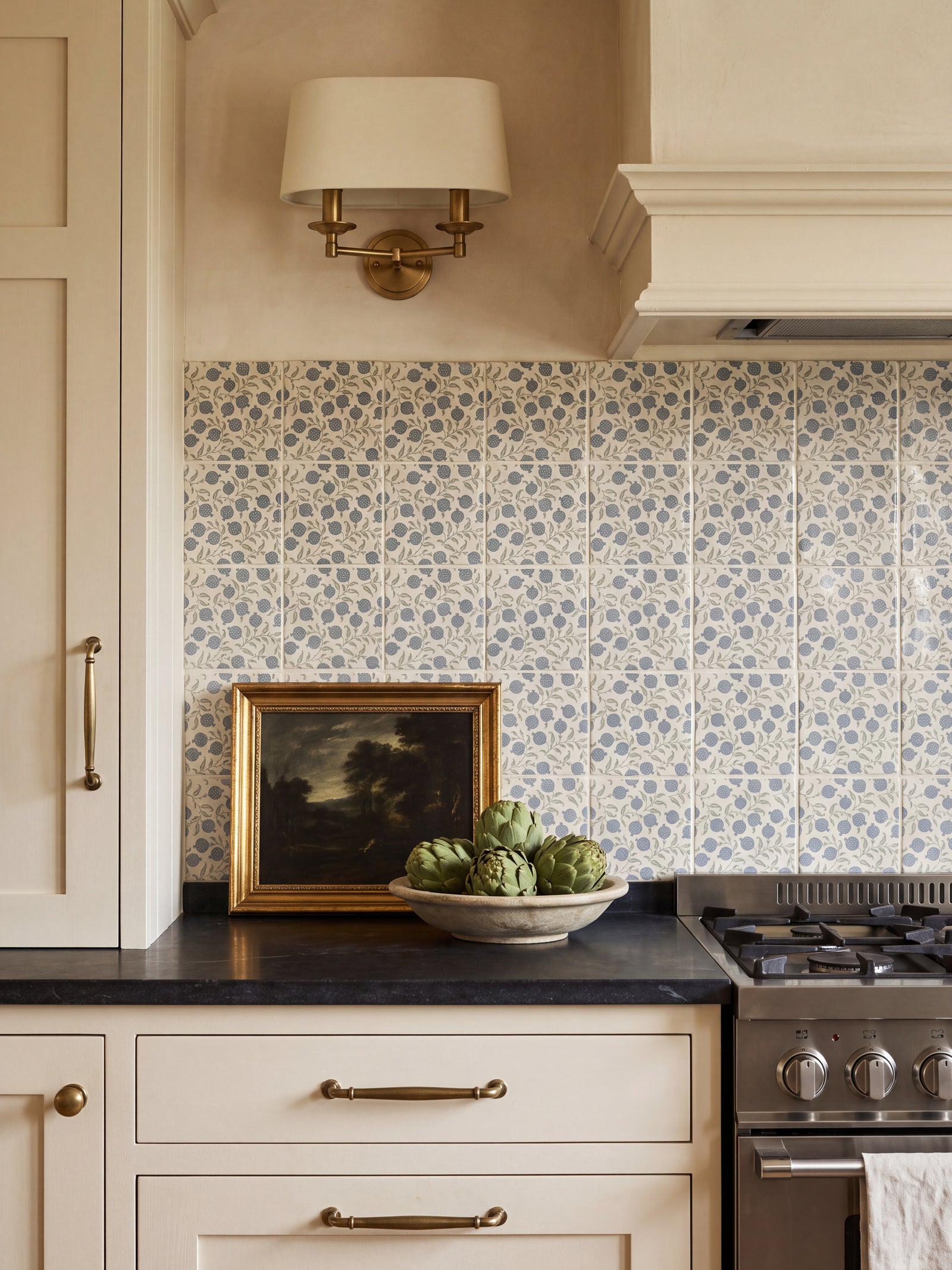 Anaar Pottery Blue tile splashback in a cream kitchen with black worktop, brass sconce and gilt-framed painting