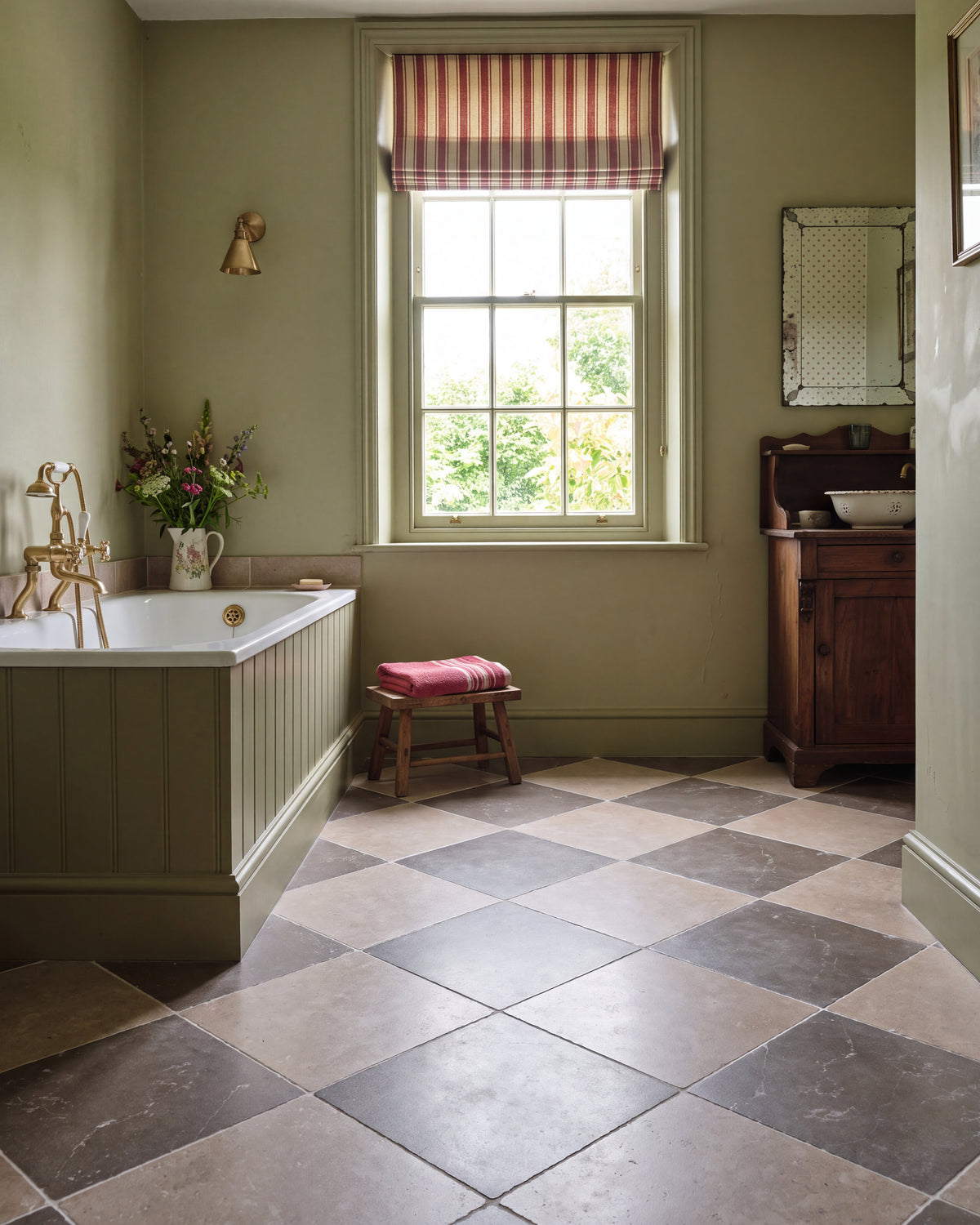 Amaya Limestone Tumbled Finish laid as a diagonal chequerboard with Wychwood Limestone Tumbled Finish in a sage green bathroom with a panelled bath, brass taps and a sash window
