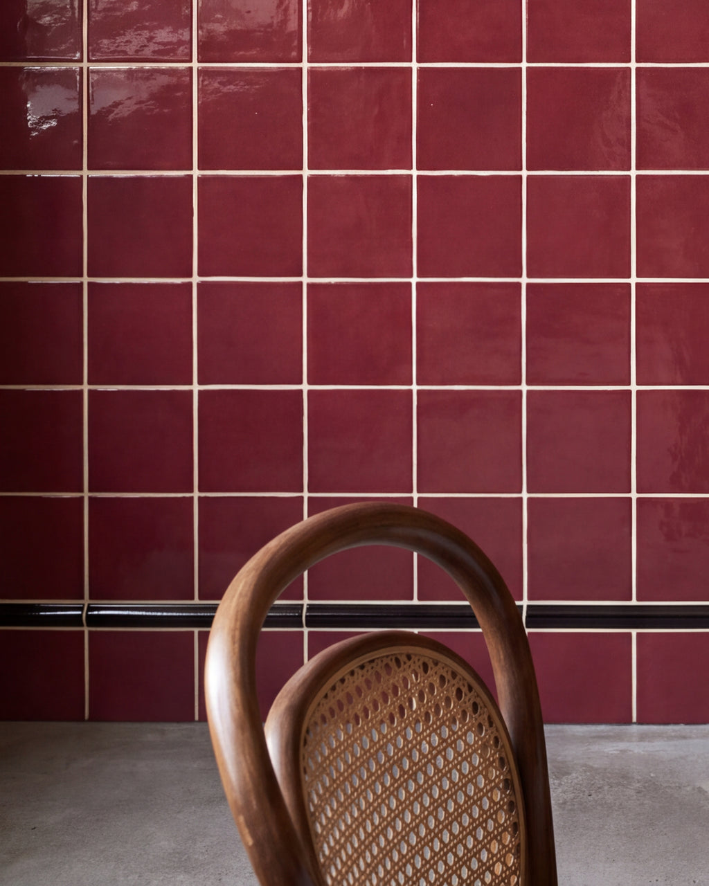 Close crop of Architectural Mouldings Bead Ceramic Black at the foot of a burgundy gloss square tiled wall, with a bentwood chair and polished concrete floor