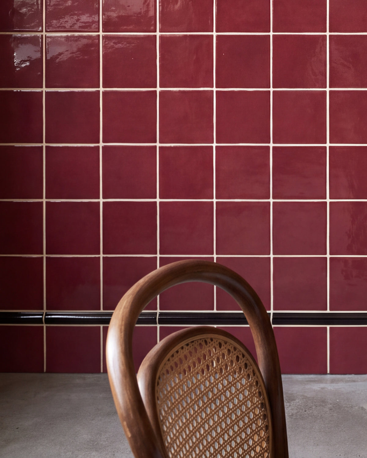 Close crop of Architectural Mouldings Bead Ceramic Black at the foot of a burgundy gloss square tiled wall, with a bentwood chair and polished concrete floor