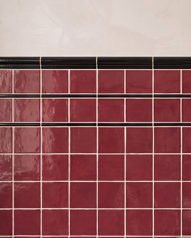 Close crop of Architectural Mouldings Dado Ceramic Black, the ribbed black rail finishing the top edge of a burgundy gloss square tiled wall against pink plaster