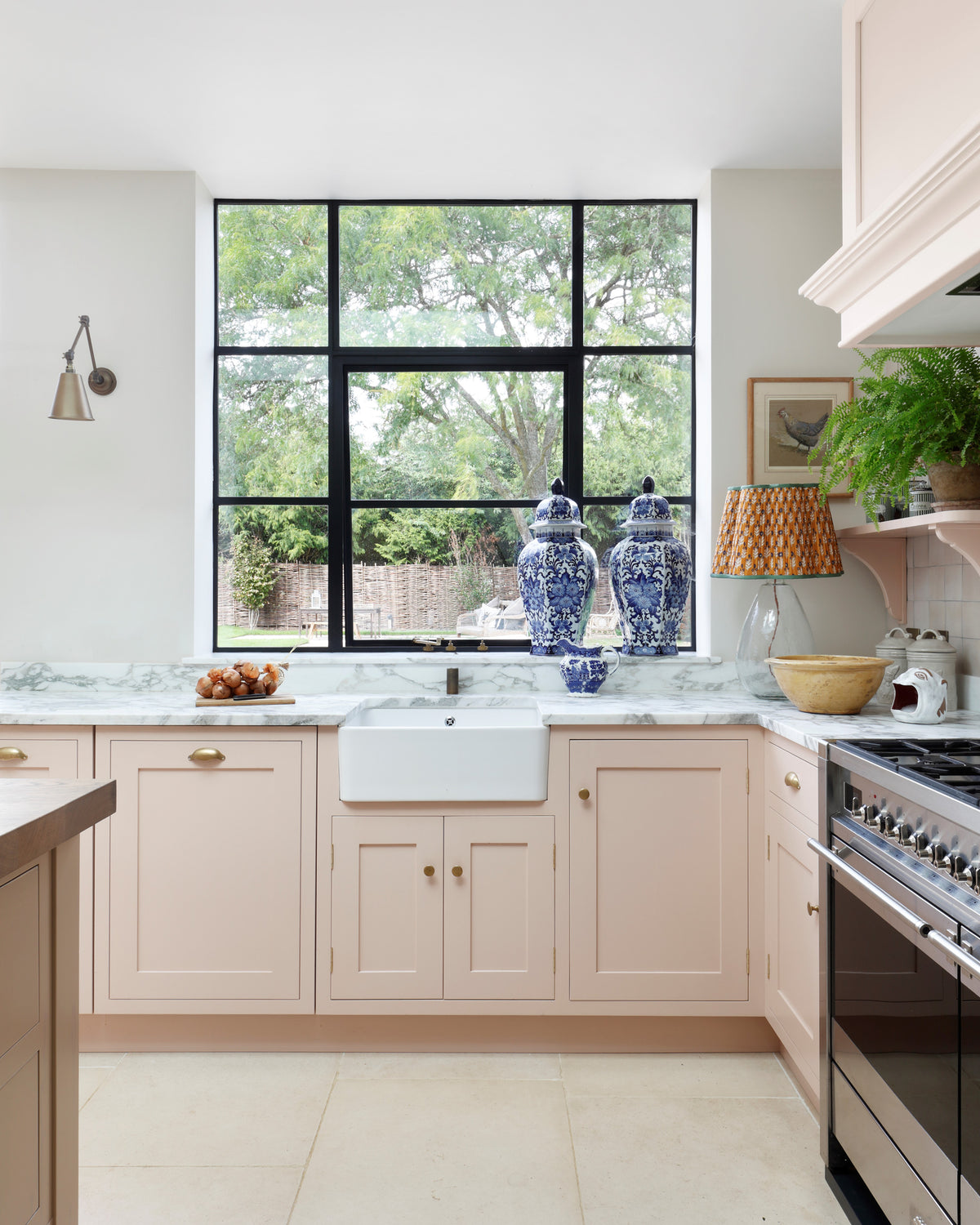 Blandford Limestone Tumbled Finish flooring in a pale pink shaker kitchen with a Belfast sink, marble worktops and a black Crittall window over the garden