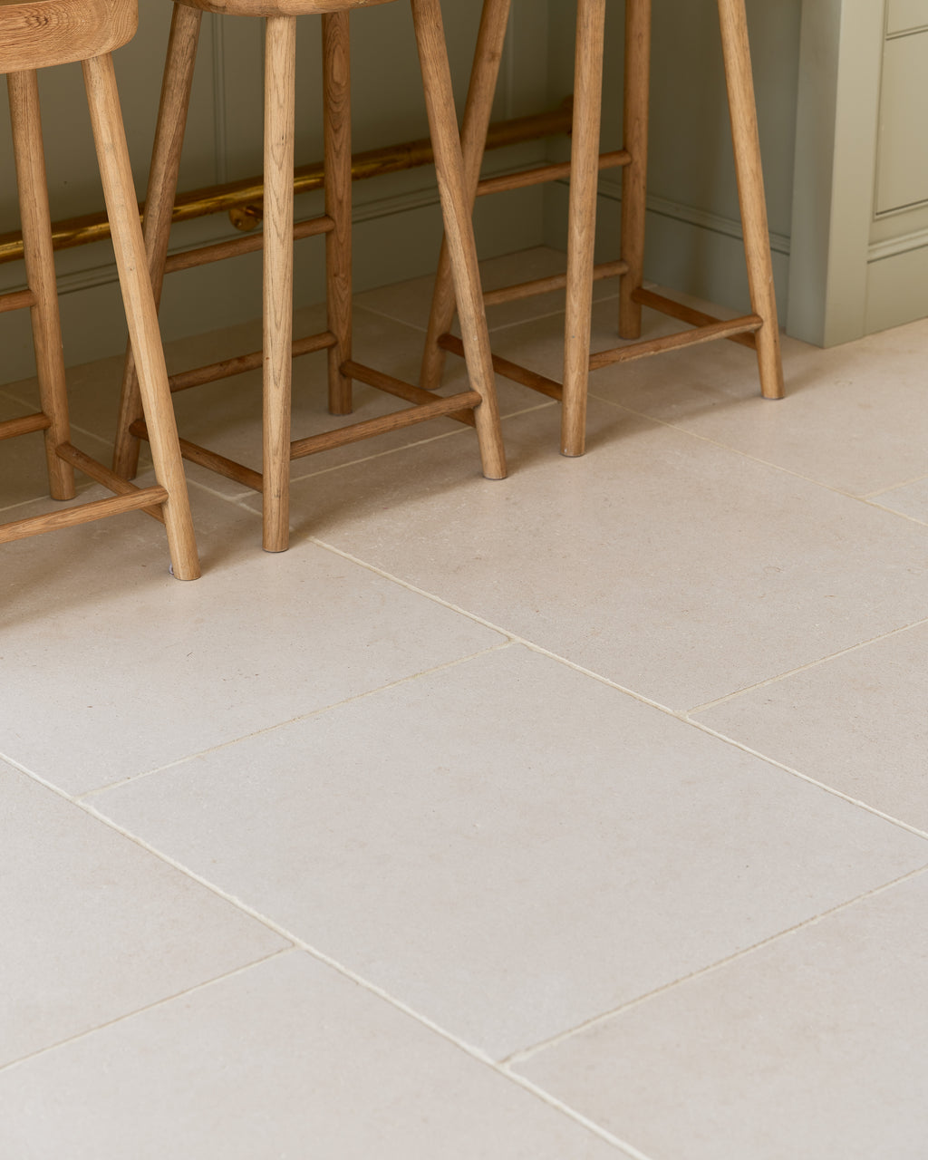 Blandford Limestone Tumbled Finish floor tiles laid square beneath a row of oak bar stools against a sage green kitchen island
