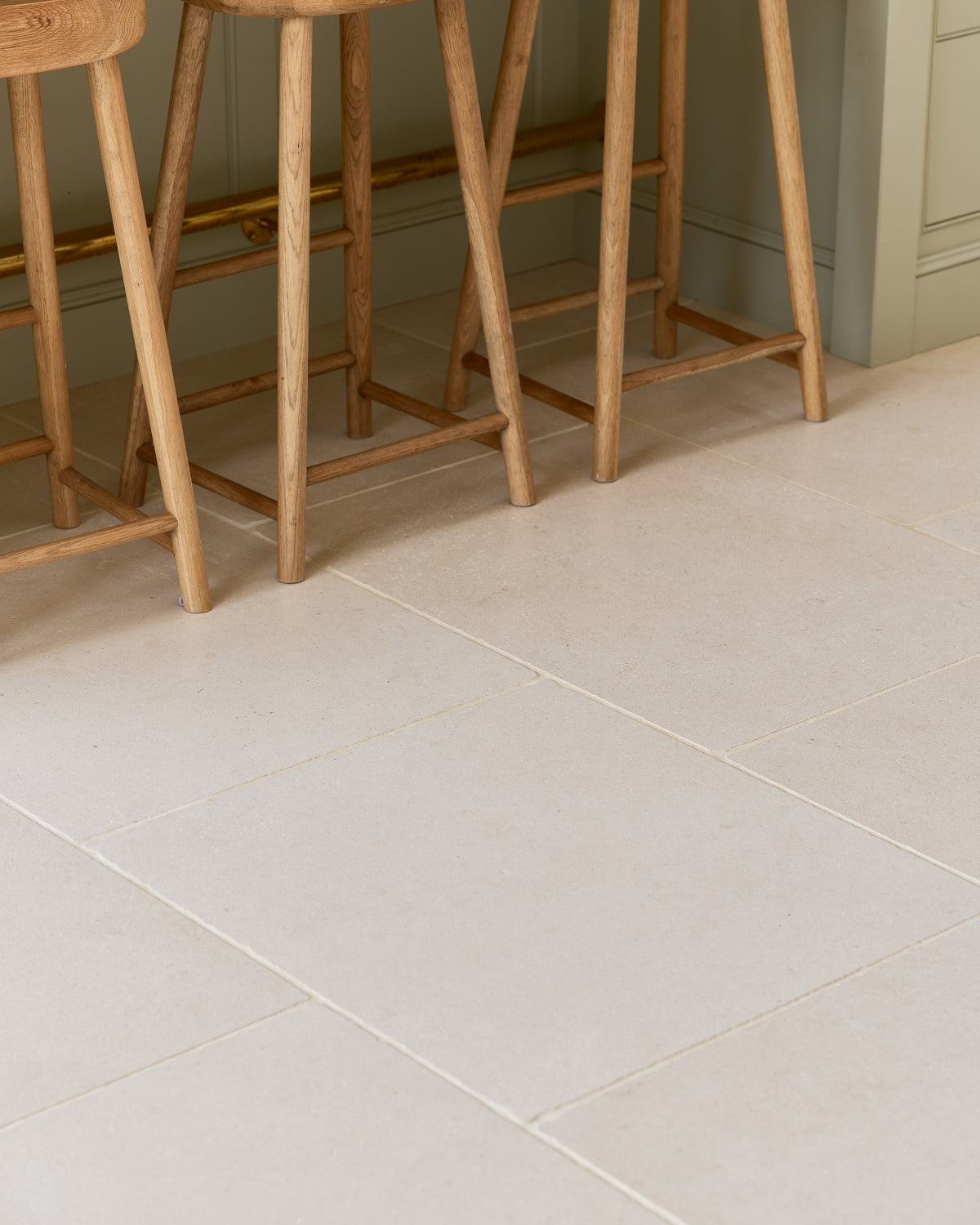 Blandford Limestone Tumbled Finish floor tiles laid square beneath a row of oak bar stools against a sage green kitchen island