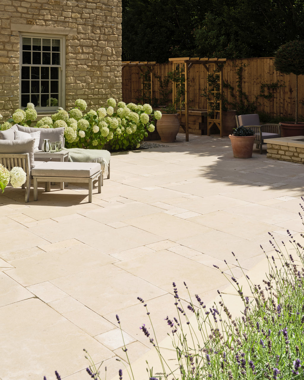 Buscot Limestone Cobble Tumbled & Etched Finish beyond a paved garden terrace with hydrangeas and day beds outside a Cotswold stone house
