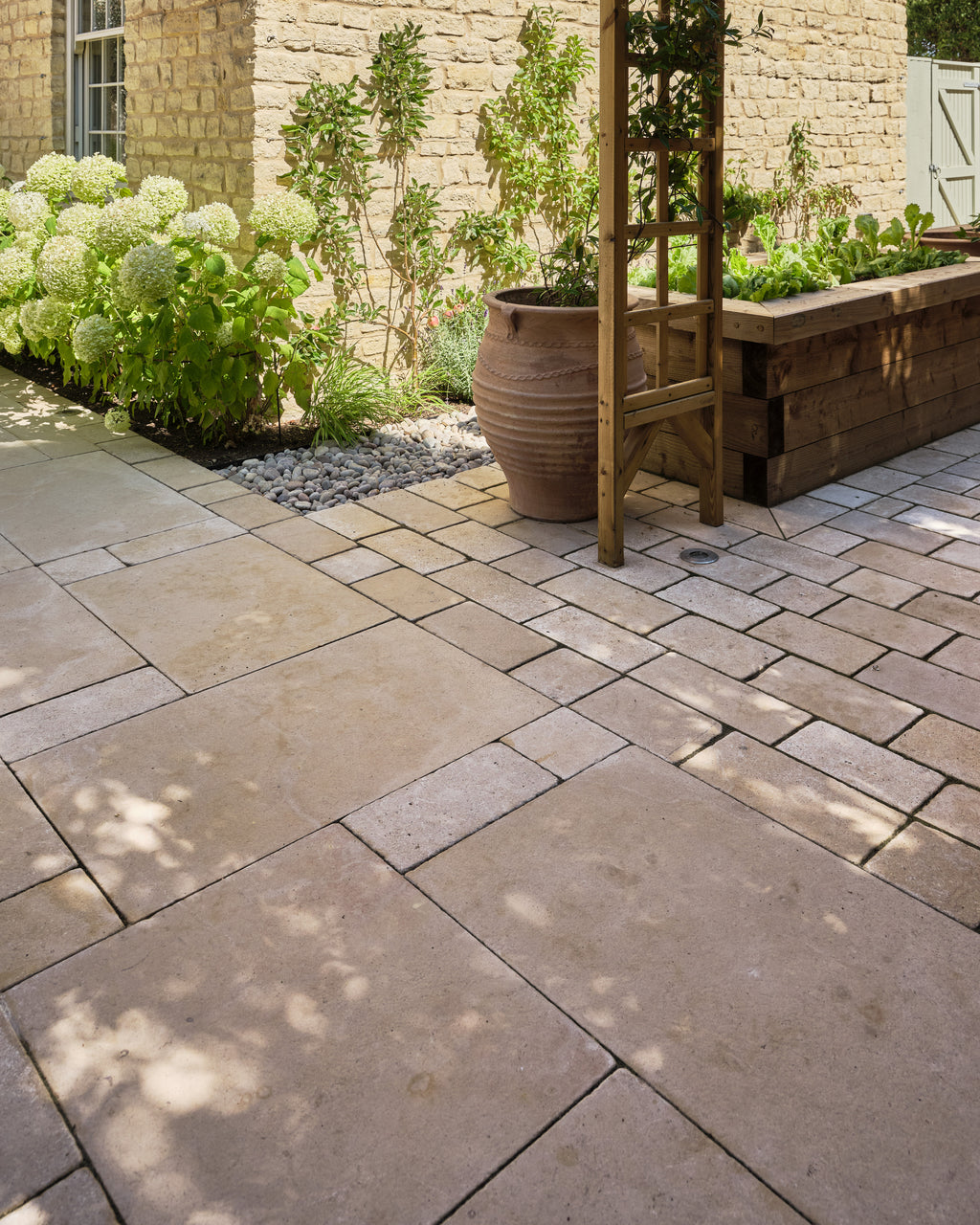 Buscot Limestone Cobble Tumbled & Etched Finish paving beside a raised timber vegetable planter and a large terracotta olive jar