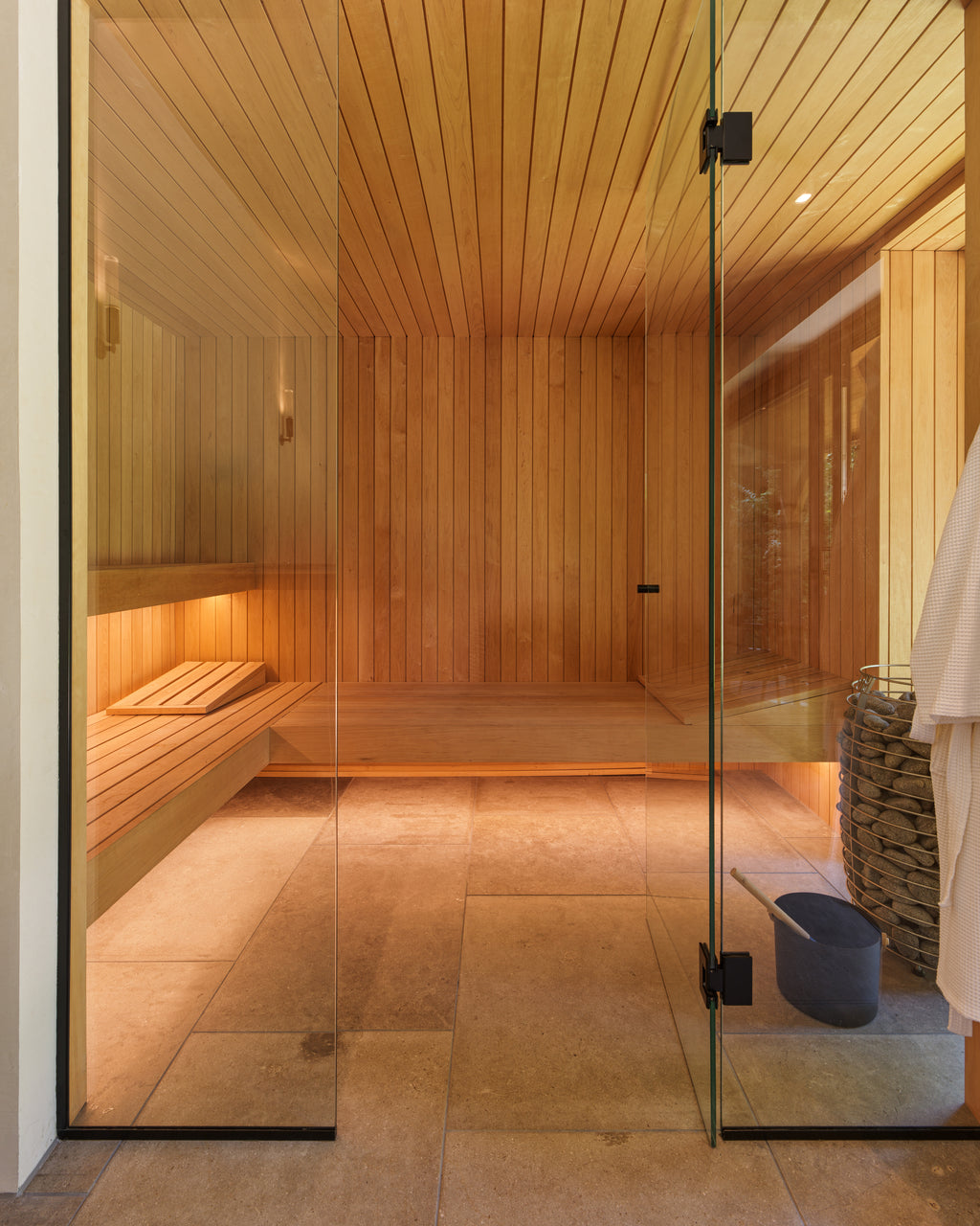 Buscot Limestone Etched Finish flooring in a cedar-lined home sauna behind a frameless glass door, with a stone-filled heater basket