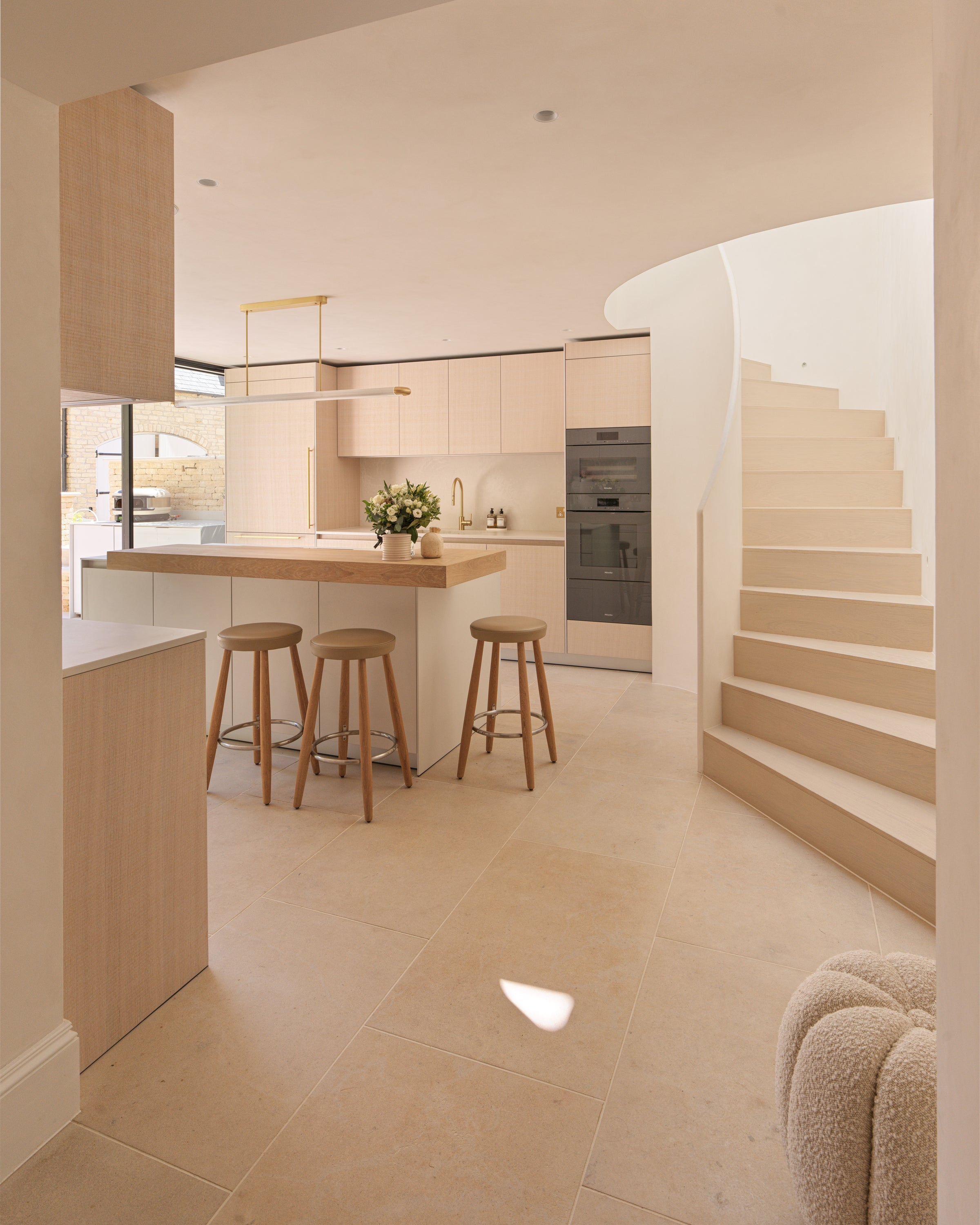 Buscot Limestone Etched Finish floor tiles running through a pale oak kitchen with an island, bar stools and a curved staircase