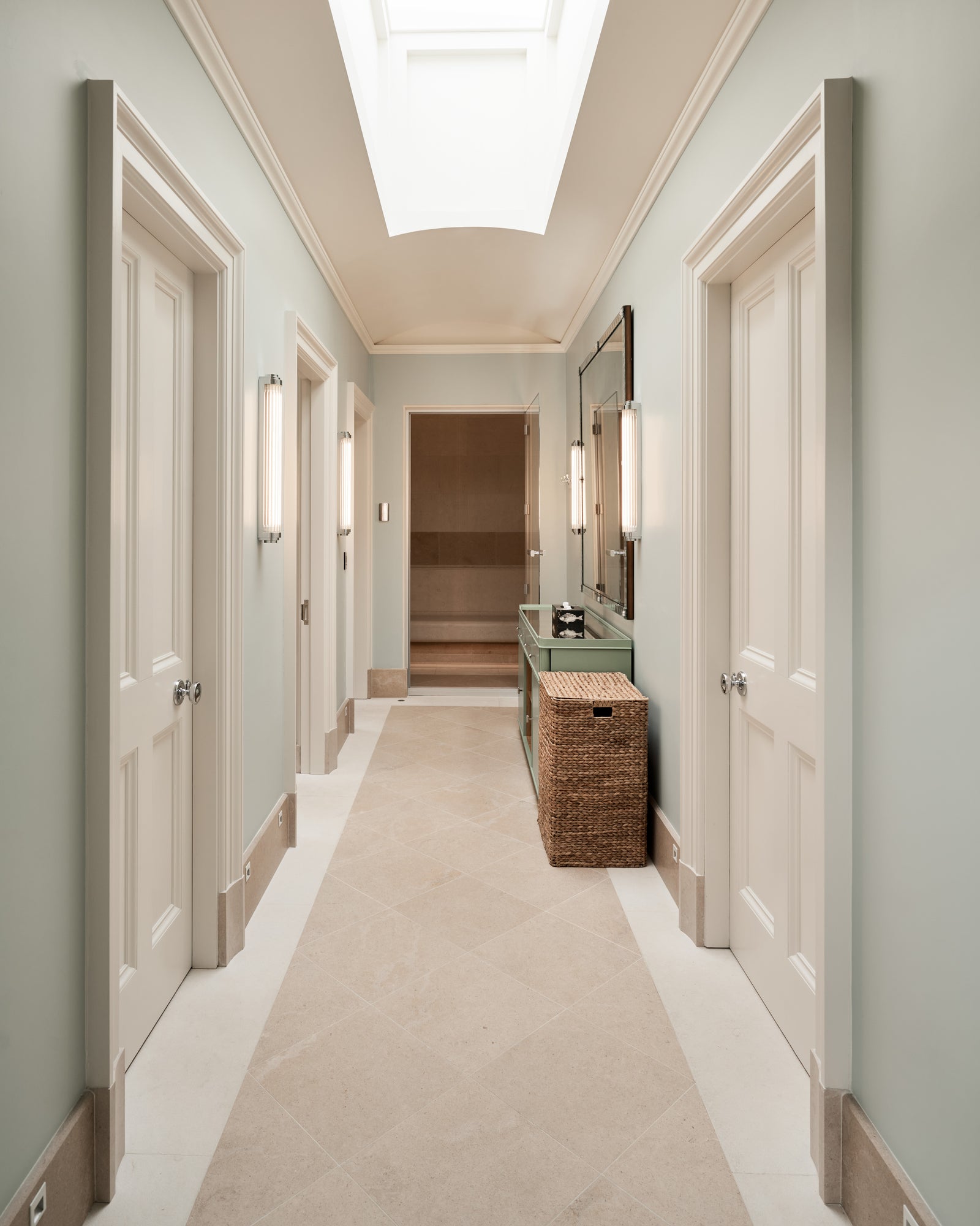 Buscot Limestone Etched Finish flooring in a skylit passage lined with panelled doors, a green console and a wicker basket