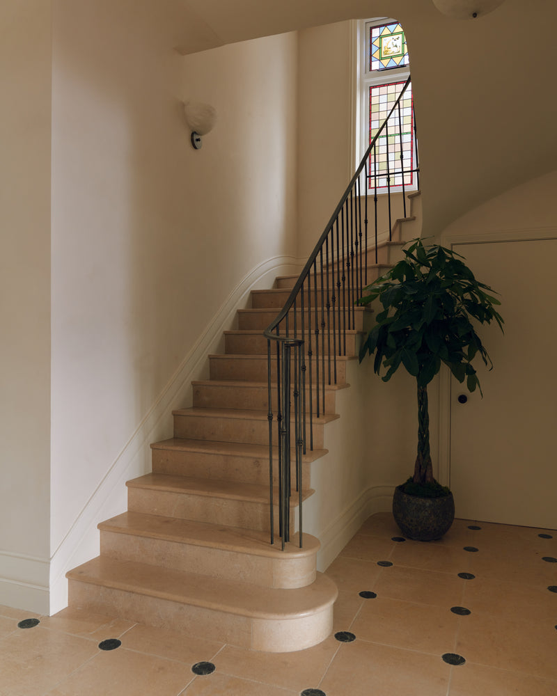 Buscot Limestone Honed Slab bespoke stone staircase with curved bullnose bottom step, wrought iron balustrade and a stained glass window above, over a limestone floor with black cabochons