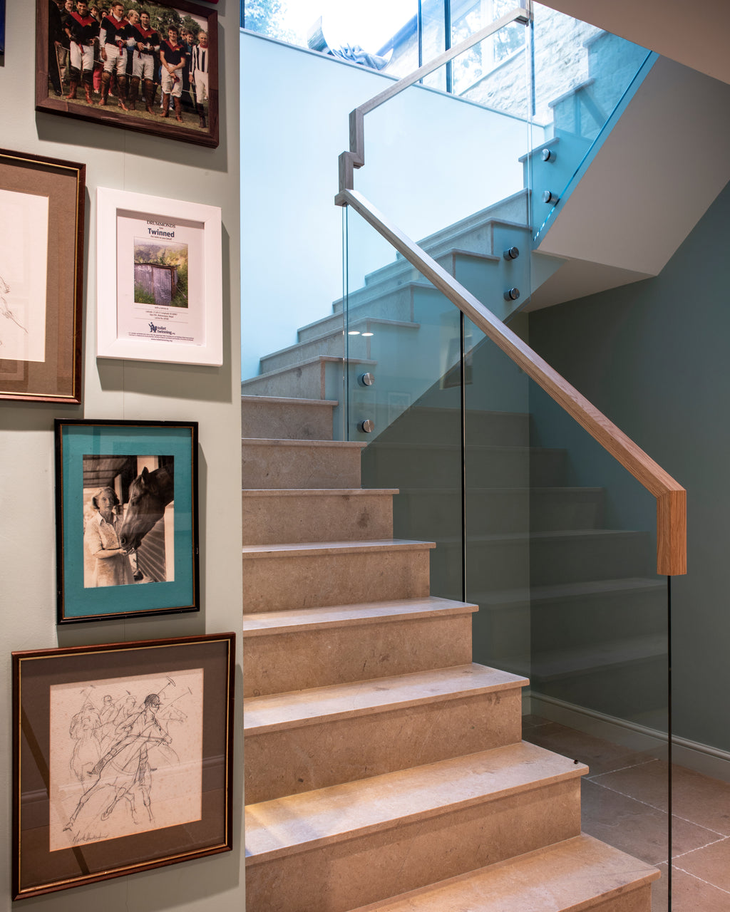 Buscot Limestone Honed Slab stair treads rising past a frameless glass balustrade with an oak handrail, beside a sage green wall hung with framed polo photographs, onto a Buscot flagged landing