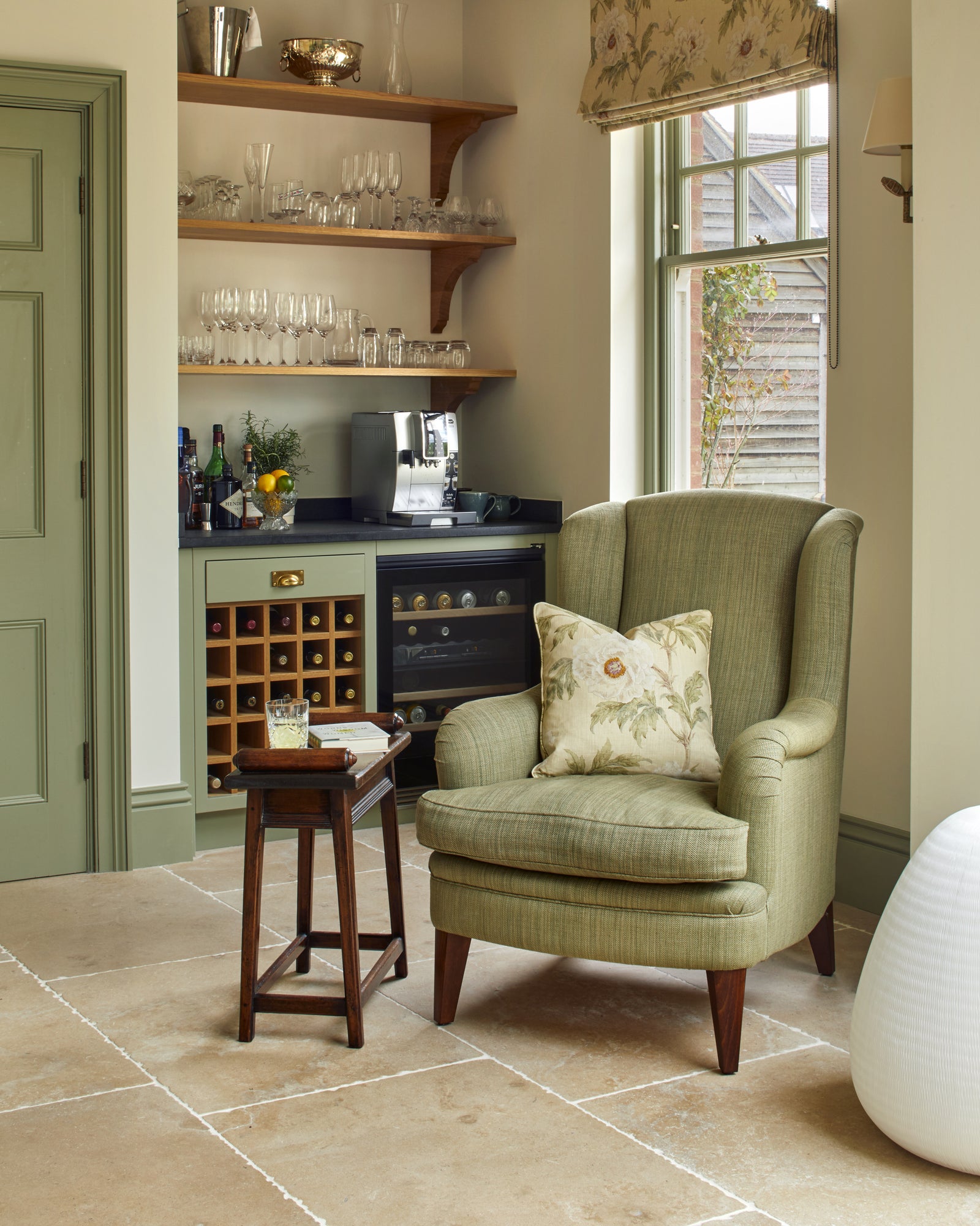 Buscot Limestone Tumbled Finish flooring beneath a wingback armchair in a green drinks nook with open shelving and a wine fridge