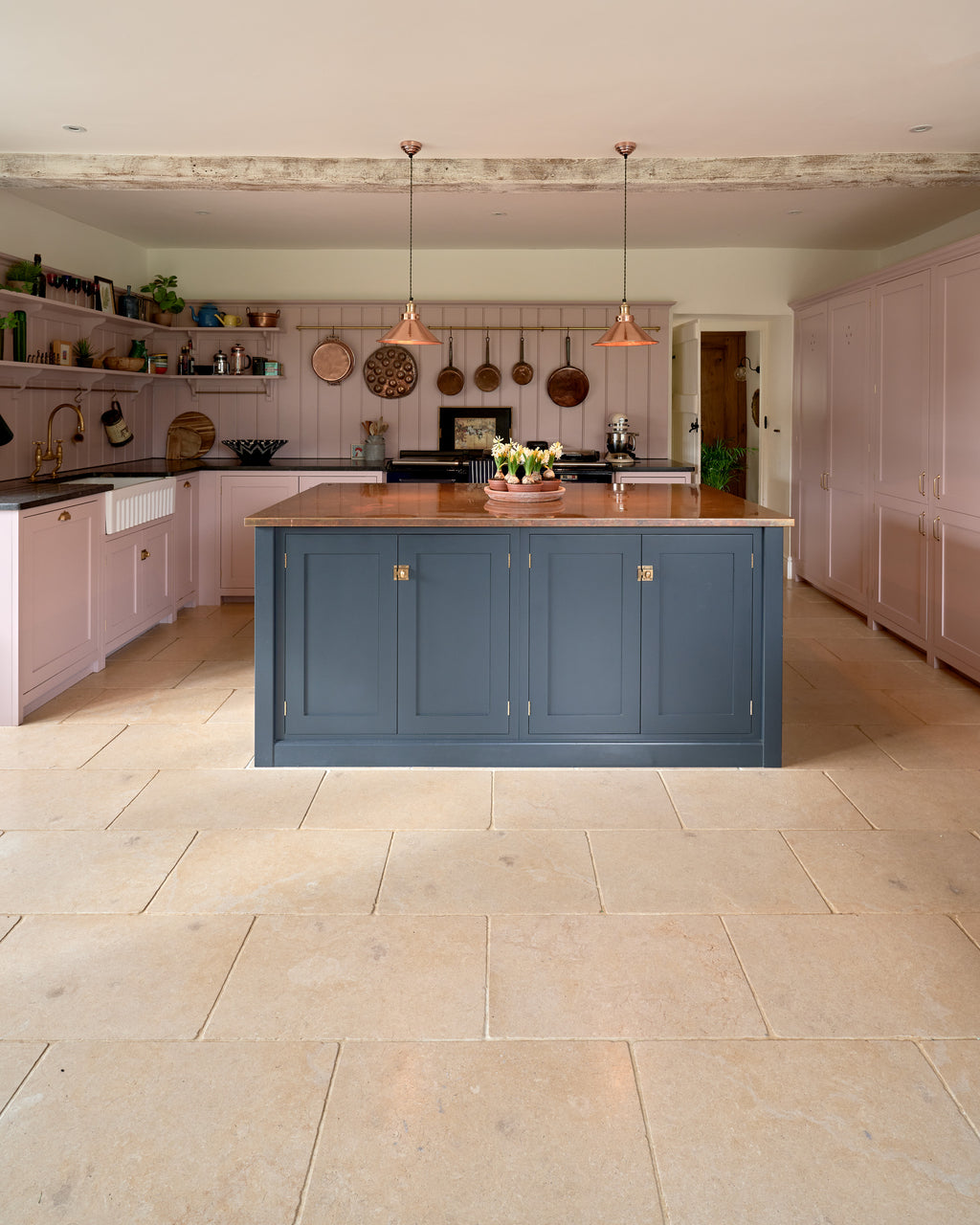 Buscot Limestone Tumbled Finish flooring in a dusky pink kitchen with a navy copper-topped island and a rail of hanging copper pans