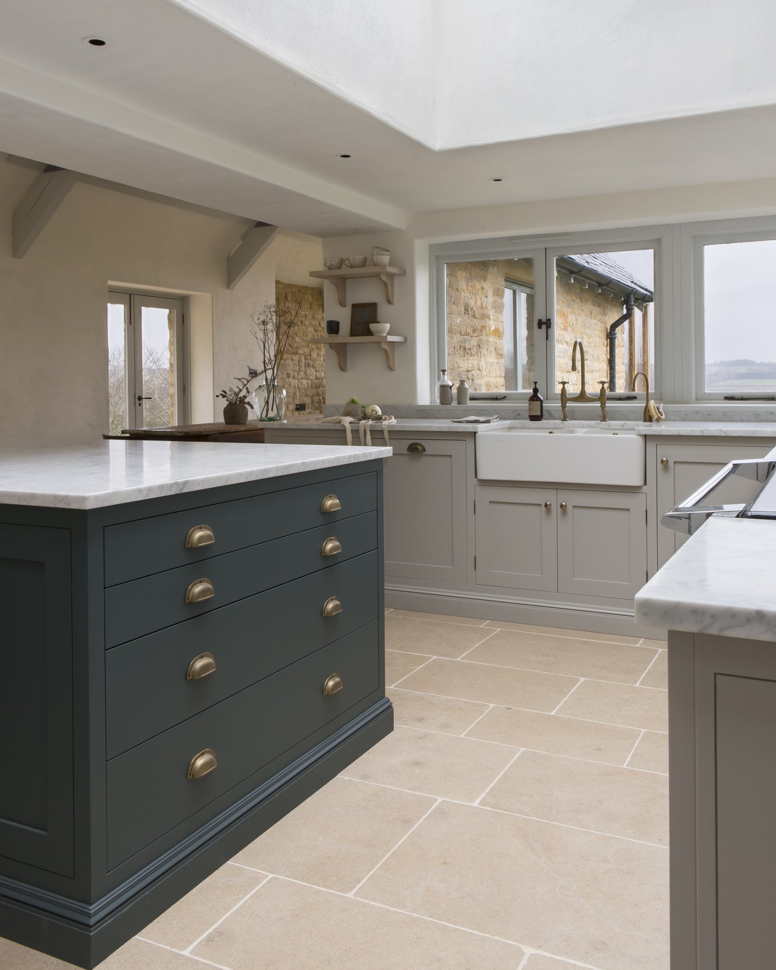 Buscot Limestone Tumbled Finish flooring in a skylit kitchen with a dark green island, marble worktops and a farmhouse sink beneath a stone-framed window