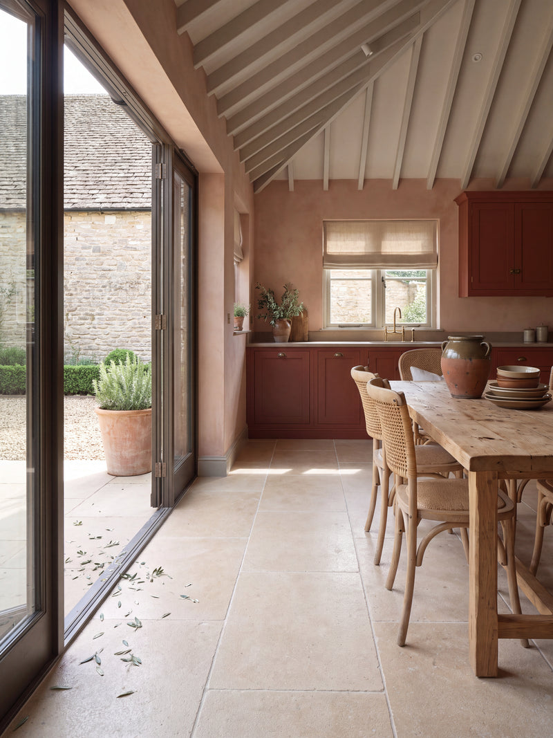 Buscot Limestone Tumbled & Etched flagstone floor in a vaulted kitchen with plaster pink walls, deep red cabinetry and bifold doors open to a Cotswold courtyard