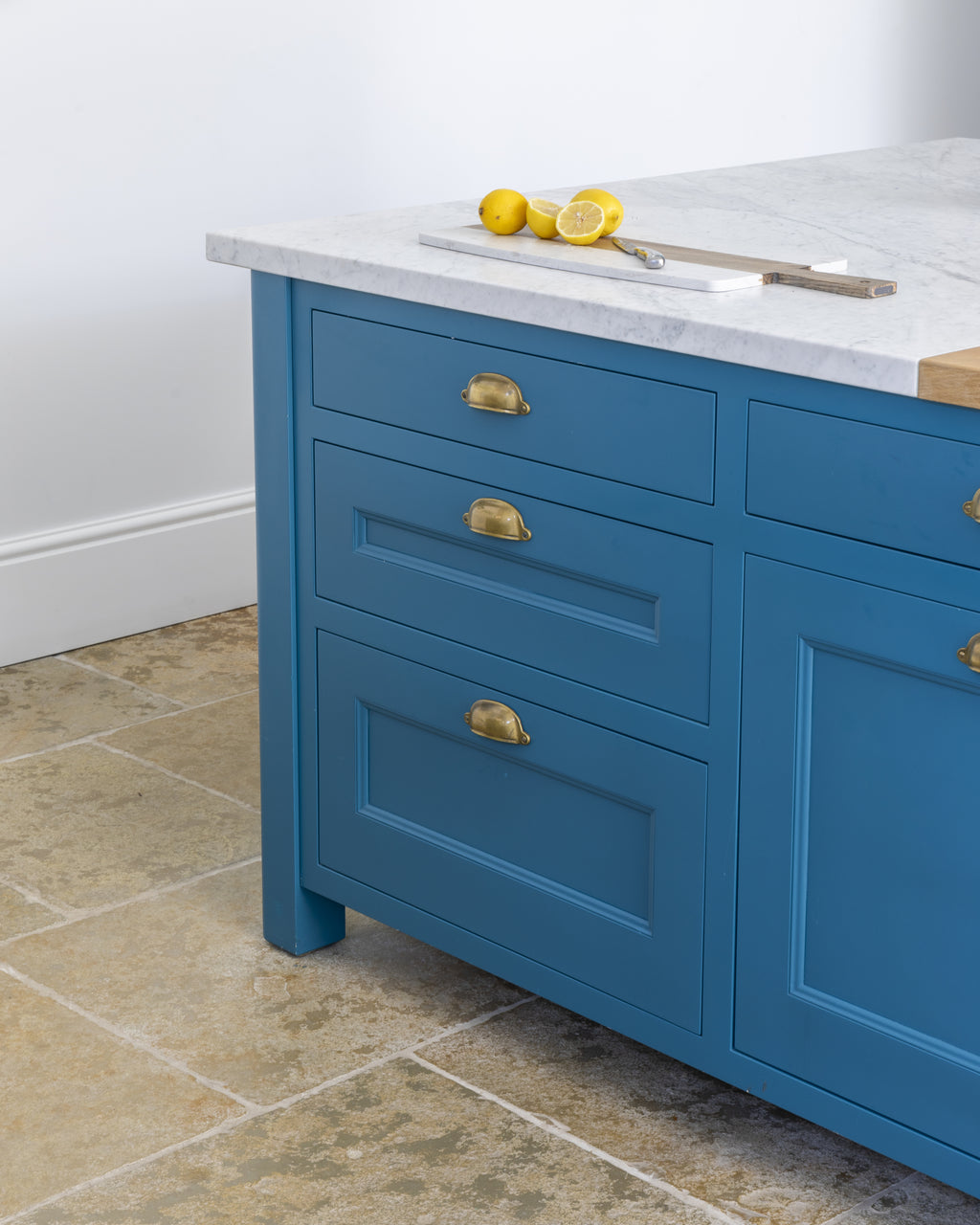 Carrara C Marble Slab as a kitchen island worktop above teal blue cabinetry with aged brass cup handles, on a tumbled limestone floor