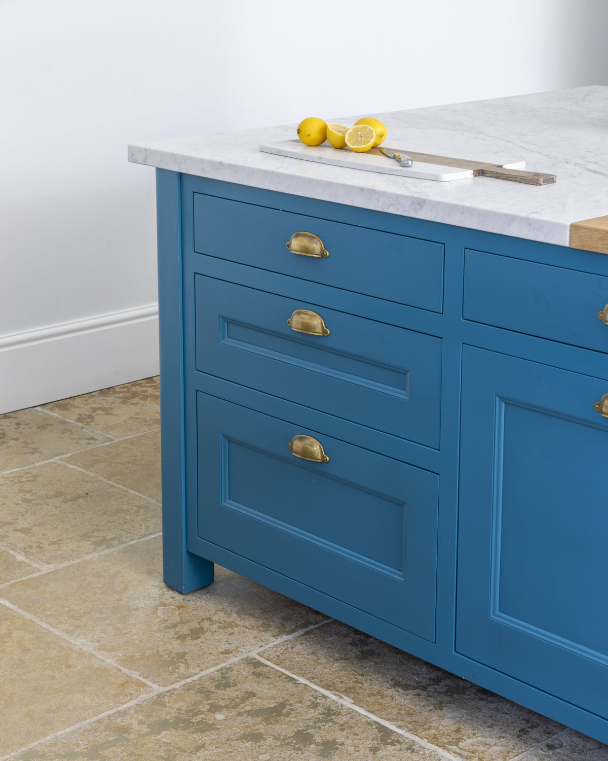 Carrara C Marble Slab as a kitchen island worktop above teal blue cabinetry with aged brass cup handles, on a tumbled limestone floor