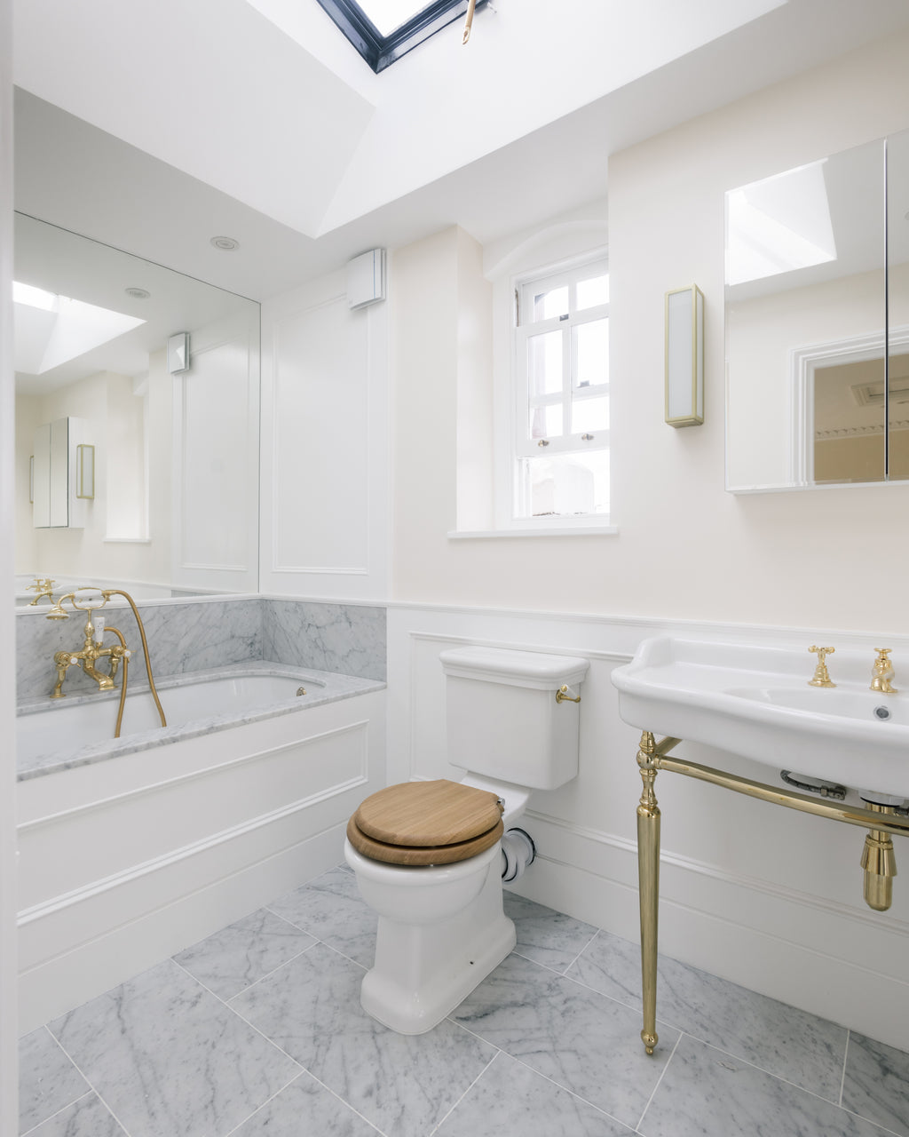 Carrara C Marble Slab forming a bath surround and deck in a white panelled bathroom beneath a pitched skylight, with a marble tiled floor and brass fittings