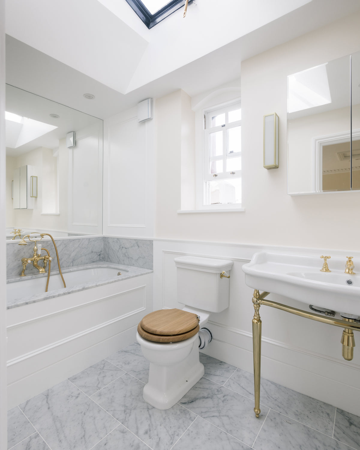 Carrara C Marble Slab forming a bath surround and deck in a white panelled bathroom beneath a pitched skylight, with a marble tiled floor and brass fittings