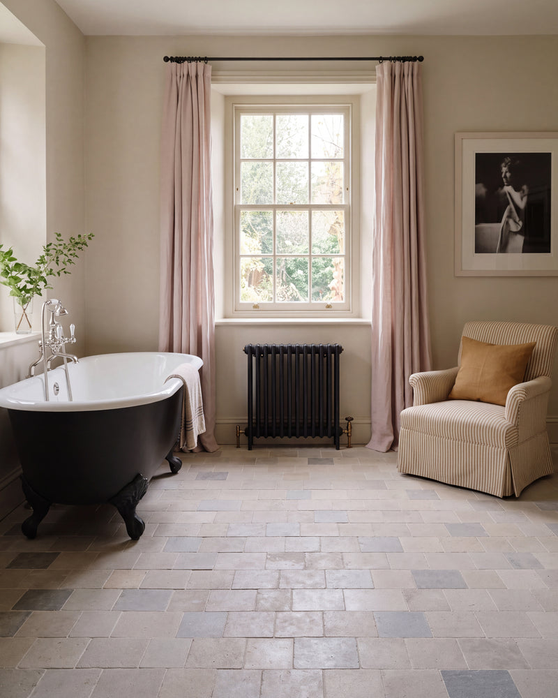 Castilan Limestone Cobbles Tumbled laid in a running bond across a bathroom floor with a black roll-top bath, cast iron radiator and ticking stripe armchair