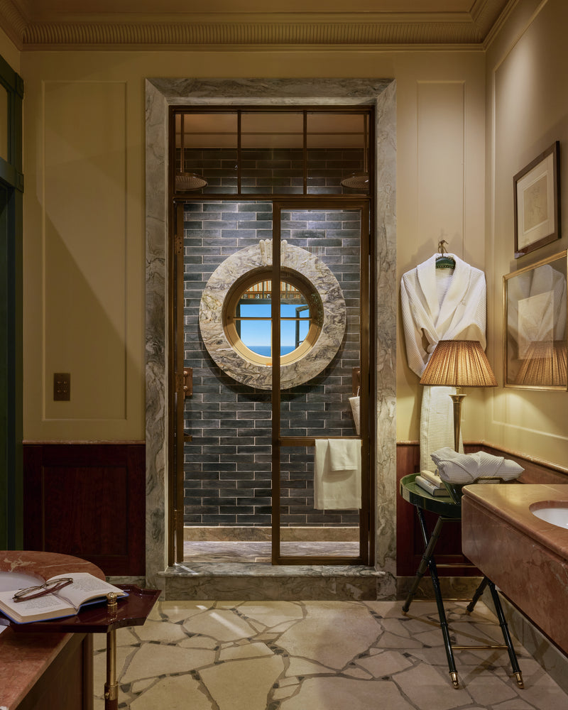 Coral Reef Quartzite Slab framing the shower doorway and porthole window in a coastal bathroom, alongside Buscot Pietra Pazza flooring, Foundry Novo Navy brick tiles and a Versailles Rouge vanity top