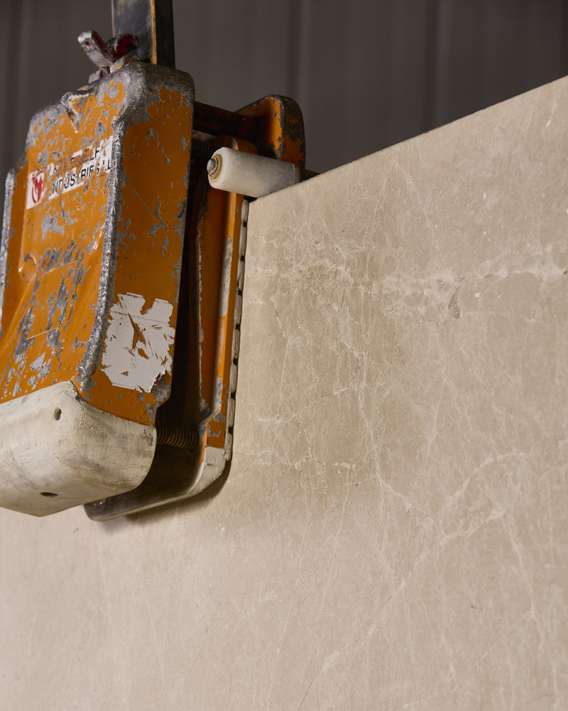 Close detail of a Domus Marble Slab edge held in a lifting clamp, showing the honed surface and fine grey veining across the cream stone