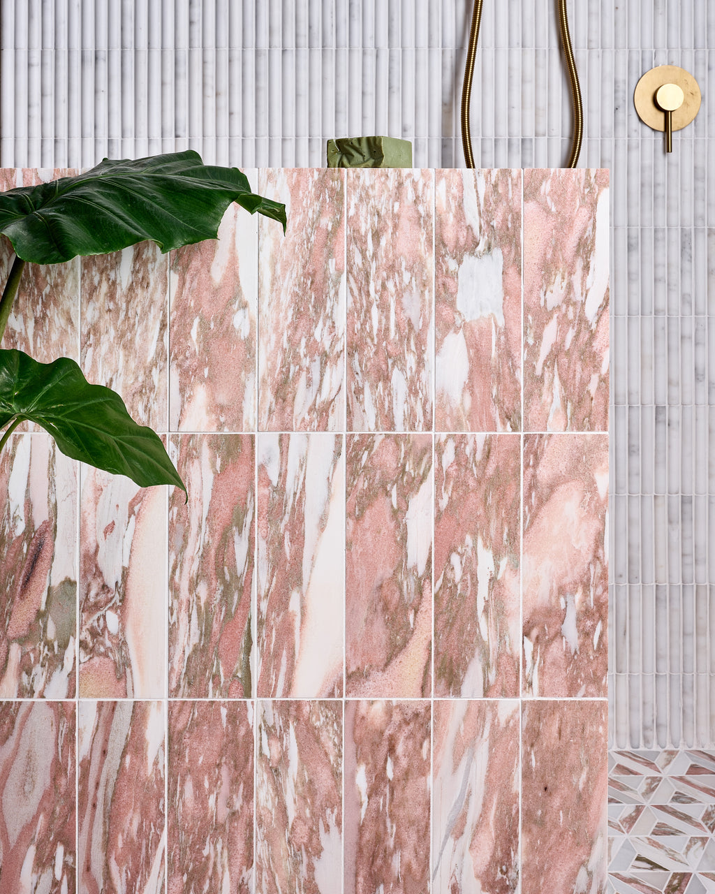 East Java Flamingo Marble Honed Brick stacked vertically across a shower wall, set against a fluted white marble mosaic, with brass shower fittings and a large-leaved houseplant in the foreground
