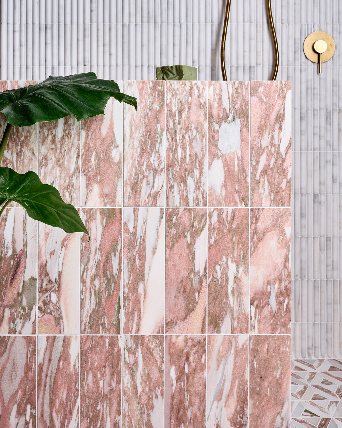 East Java Flamingo Marble Honed Brick stacked vertically across a shower wall, set against a fluted white marble mosaic, with brass shower fittings and a large-leaved houseplant in the foreground