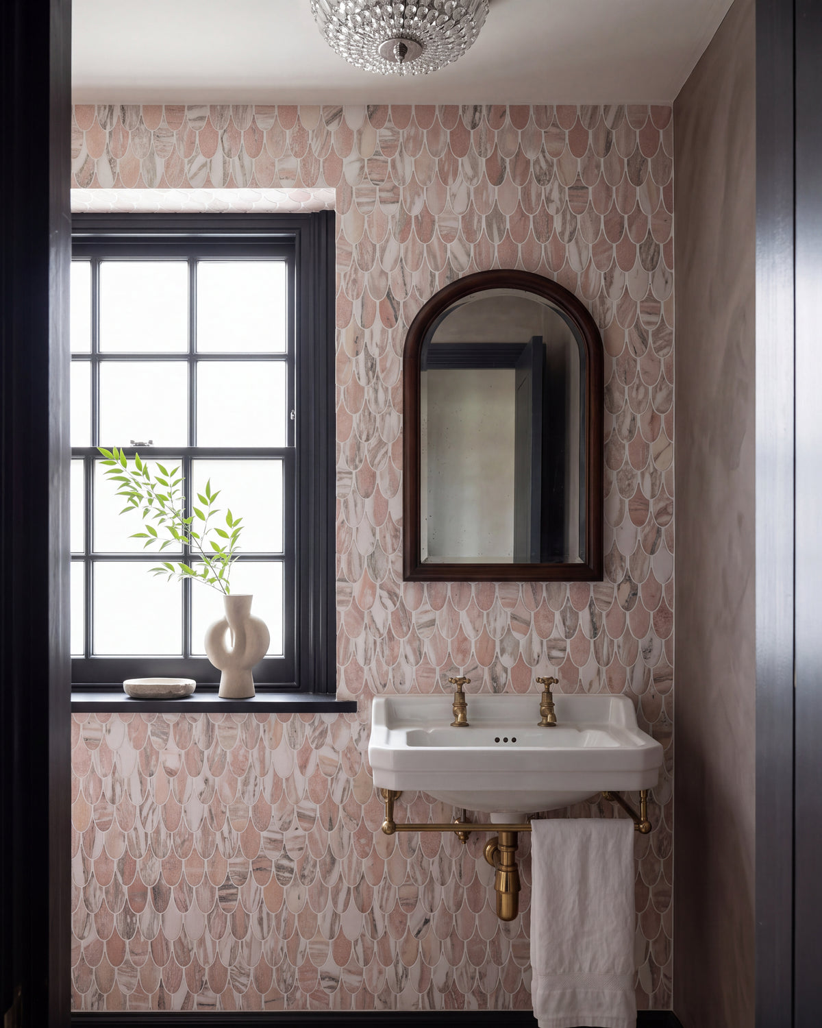 East Java Flamingo Marble Plume Mosaic running floor to ceiling in a small cloakroom with black joinery, a wall-hung basin on brass legs, an arched mirror and a crystal ceiling light