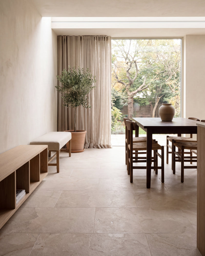 Enki Limestone Velvet Finish in large format across a pale plastered dining room, with a dark oak table and rush-seat chairs beside a full-height window onto the garden