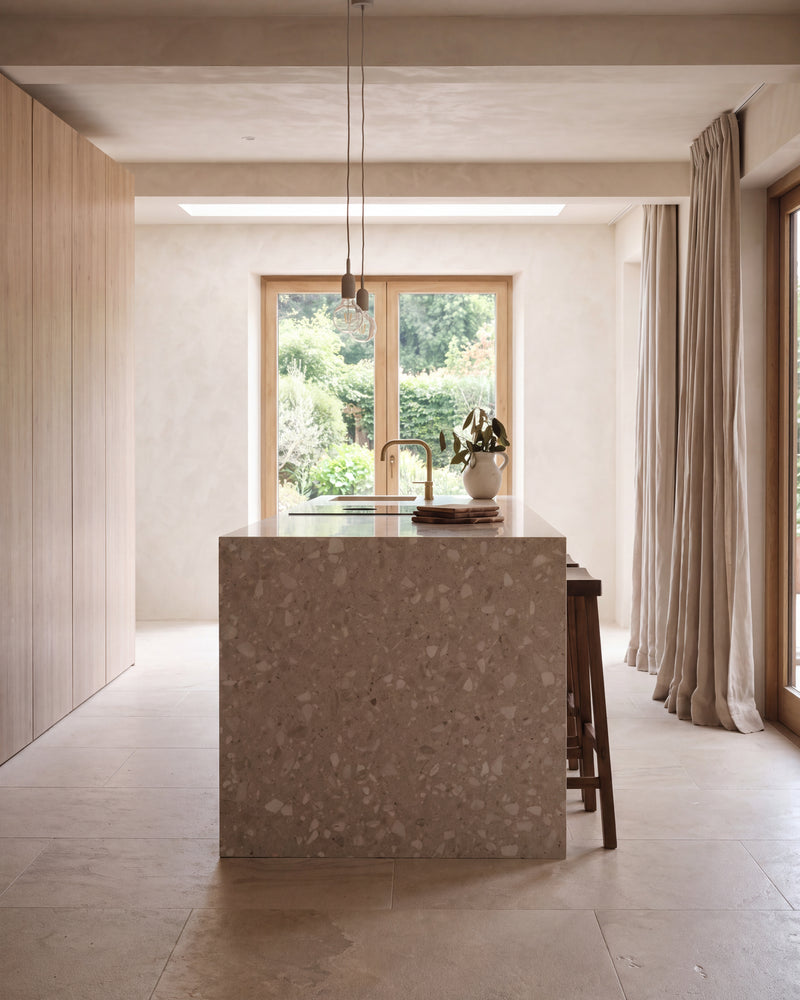 Enki Limestone Velvet Finish in large format around a waterfall terrazzo kitchen island, lit by a rooflight and a garden window with a brass tap