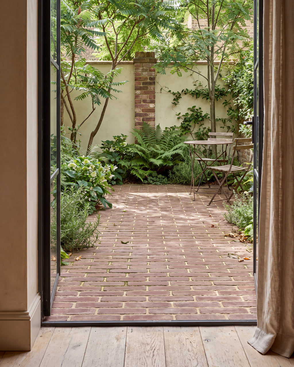 Flanders Warm Russet Tumbled Clay Pavers laid in stretcher bond across a small walled courtyard with a bistro table, seen through open steel-framed doors