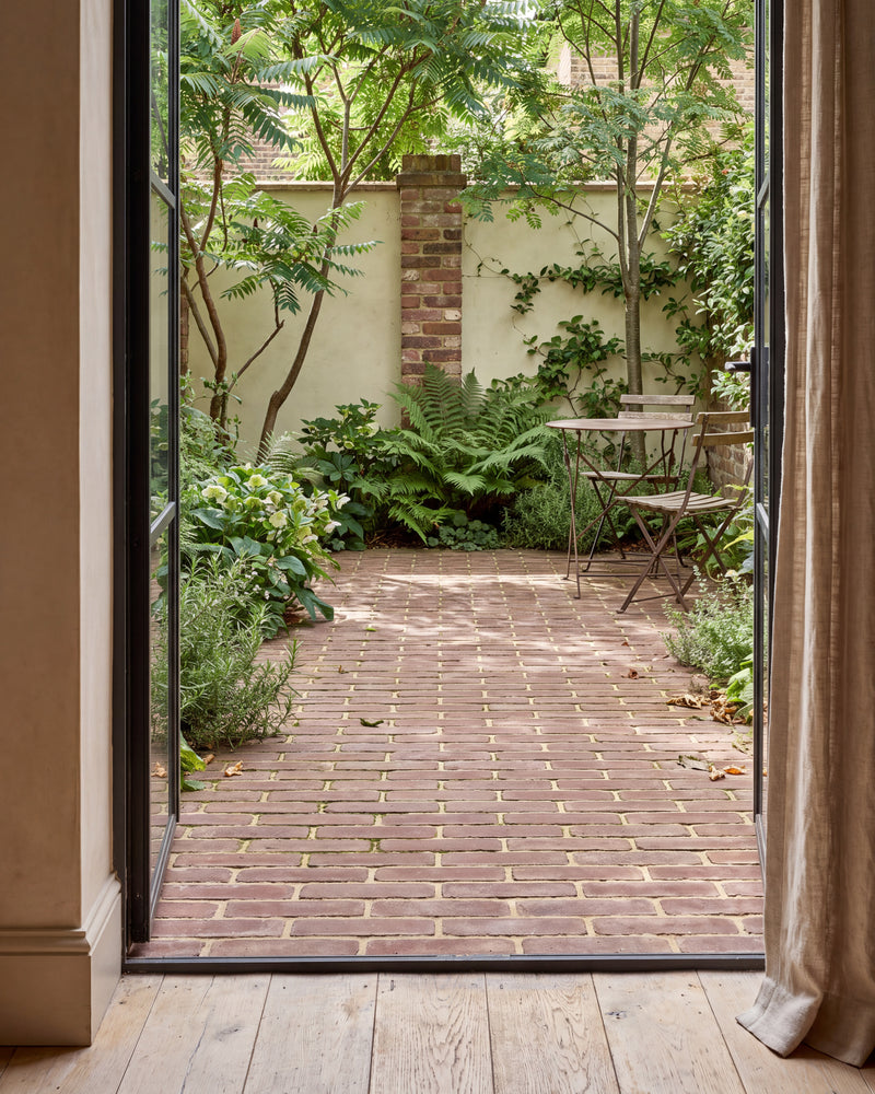 Flanders Warm Russet Tumbled Clay Pavers laid in stretcher bond across a small walled courtyard with a bistro table, seen through open steel-framed doors