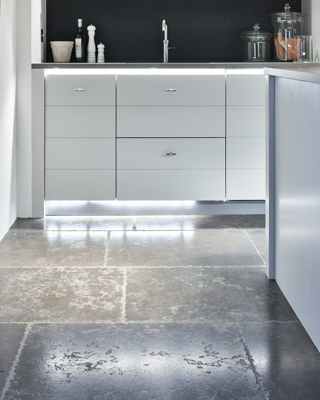 Hazlebury Limestone Seasoned Finish flagstones catching low light in a handleless white kitchen with a dark splashback