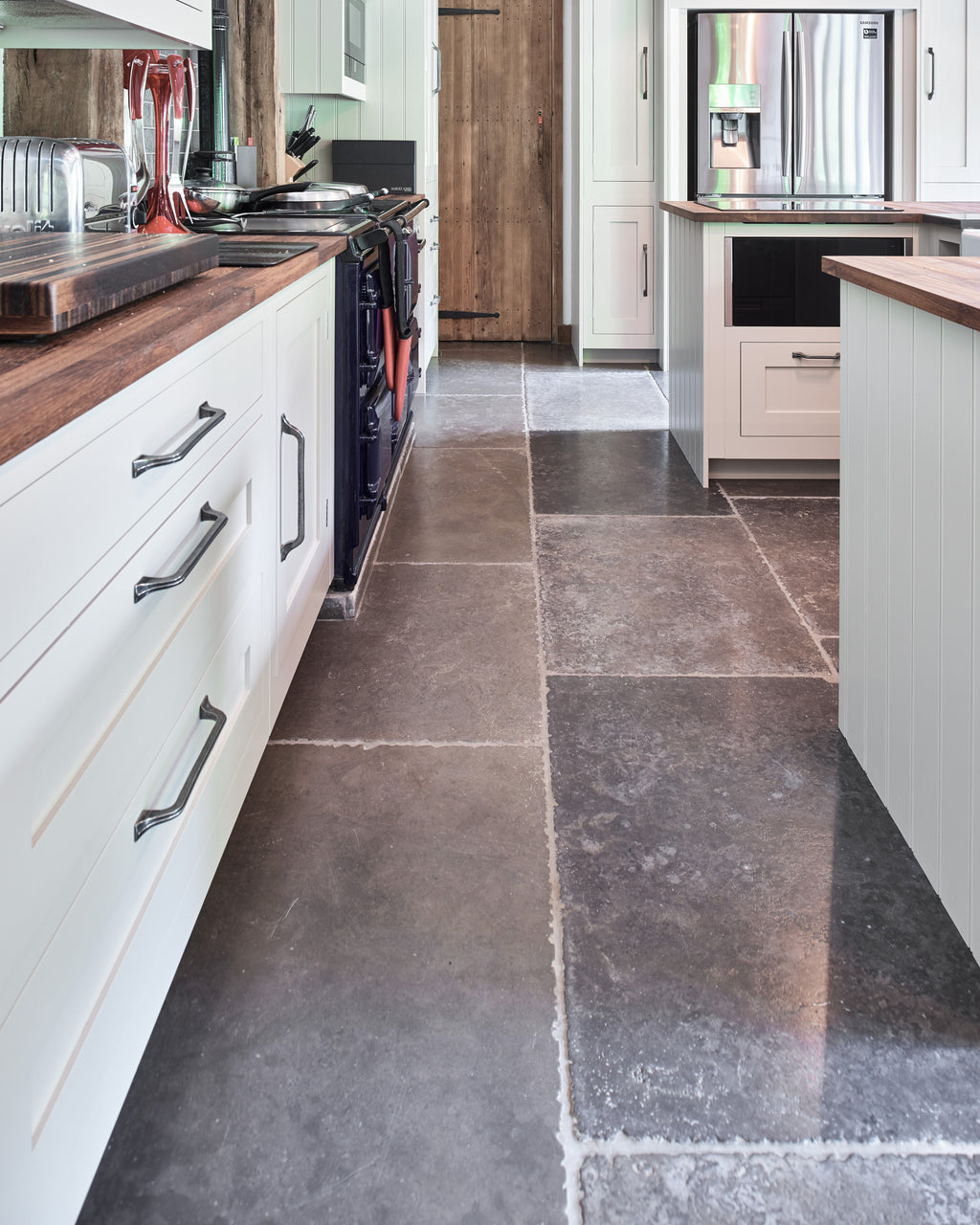 Hazlebury Limestone Seasoned Finish running the length of a narrow barn kitchen between painted cabinetry and a navy range cooker