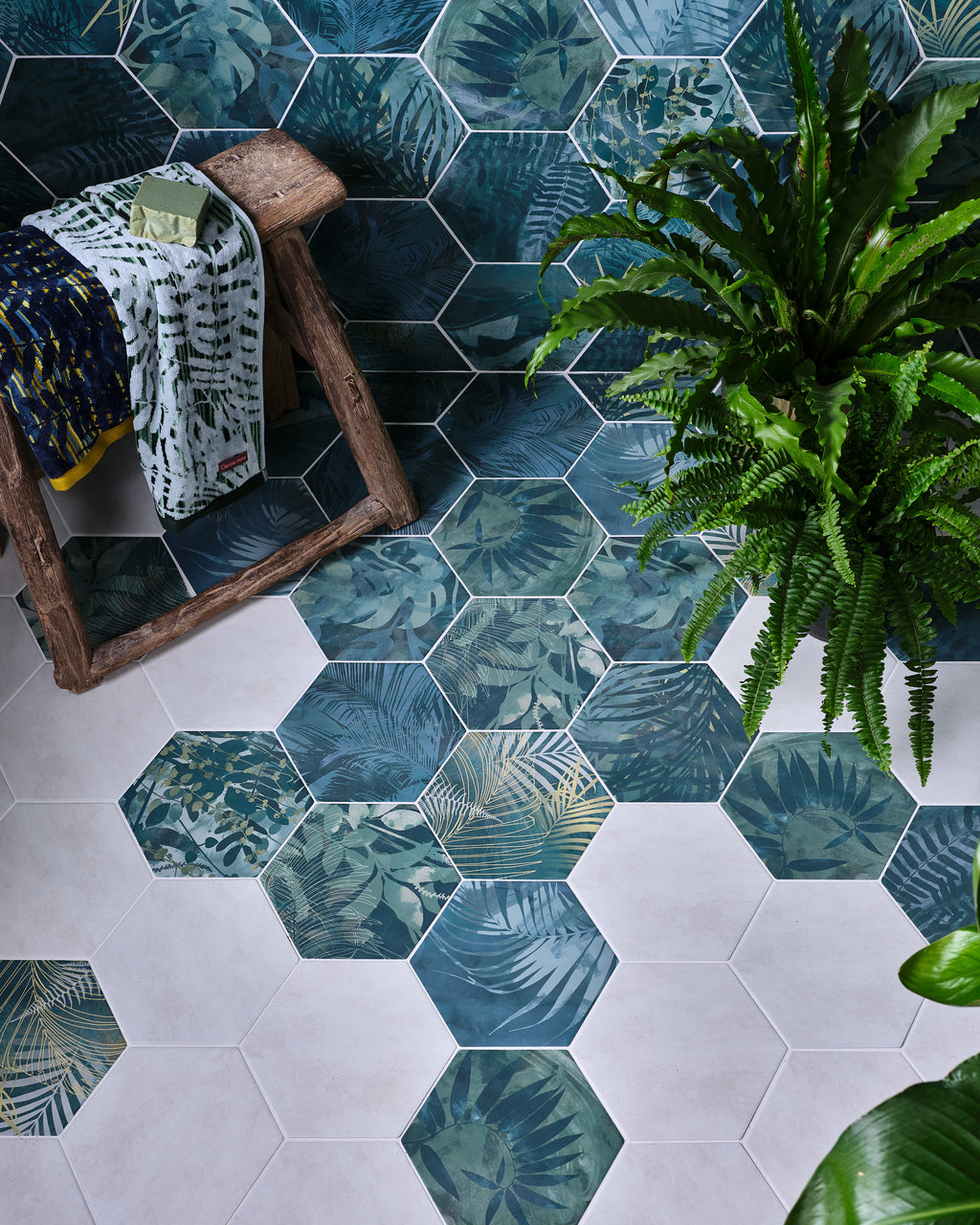 Jungle Porcelain by Clarissa Hulse hexagons scattered across a bathroom floor, the teal leaf pattern dissolving into plain pale hexagons beside a rustic stool and ferns