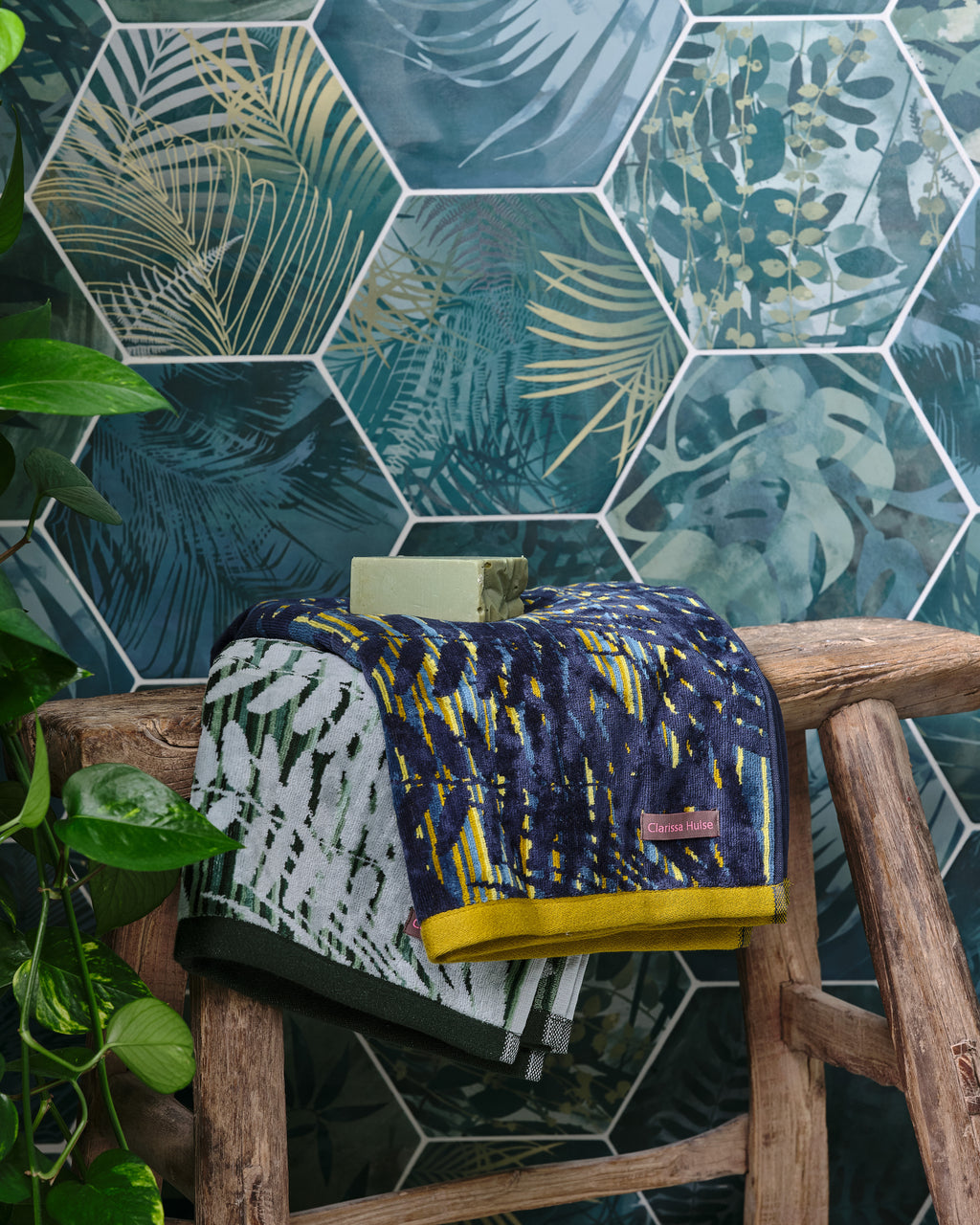 Close-up of Jungle Porcelain by Clarissa Hulse hexagons in teal and gold palm print, behind folded Clarissa Hulse towels and a bar of olive soap on a rustic wooden stool