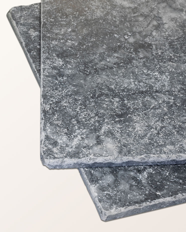 Two Kendal Marble Tumbled Finish tiles stacked at an angle against a plain background, showing the dark grey surface and chipped tumbled edges