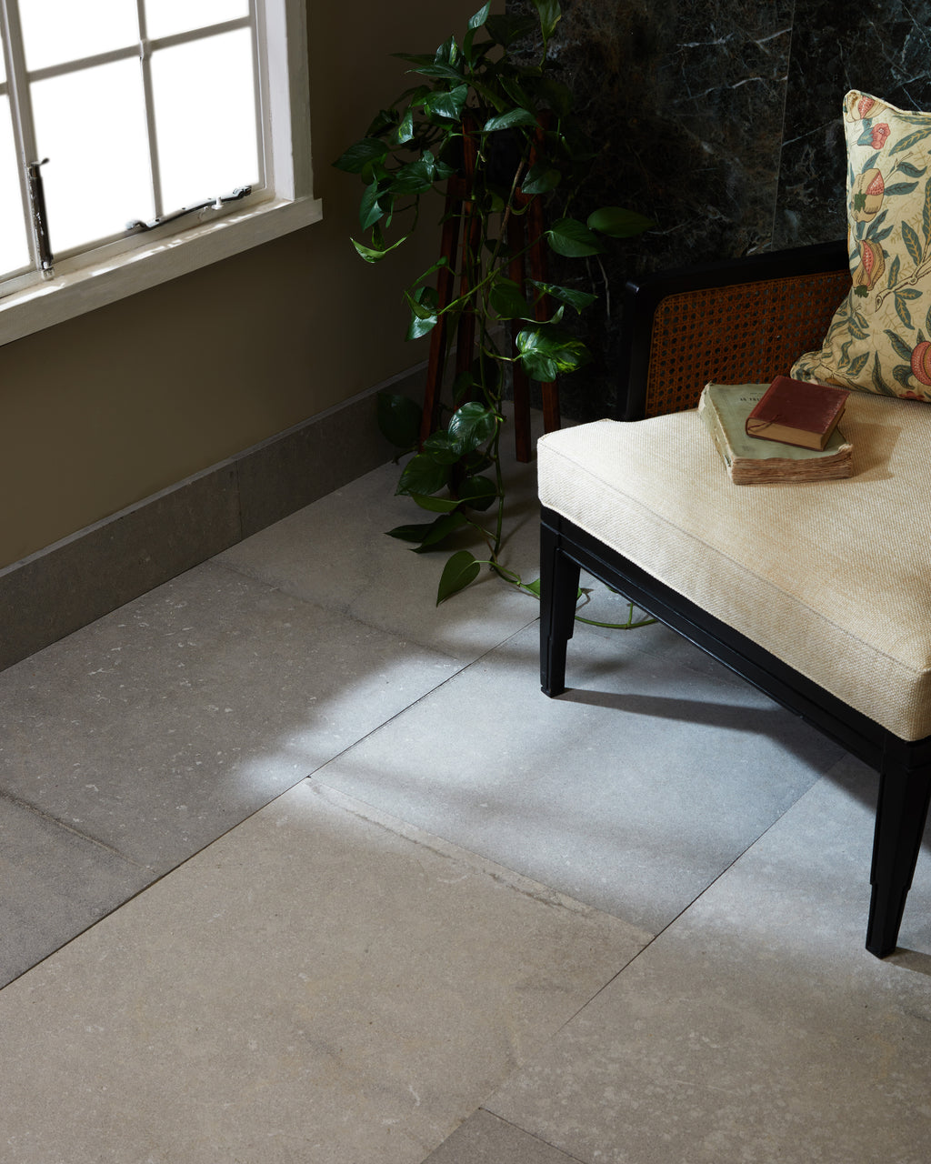 Kimmeridge Limestone Etched Finish in large format underfoot, catching low sunlight beside a black cane bench, trailing pothos and a dark green marble wall