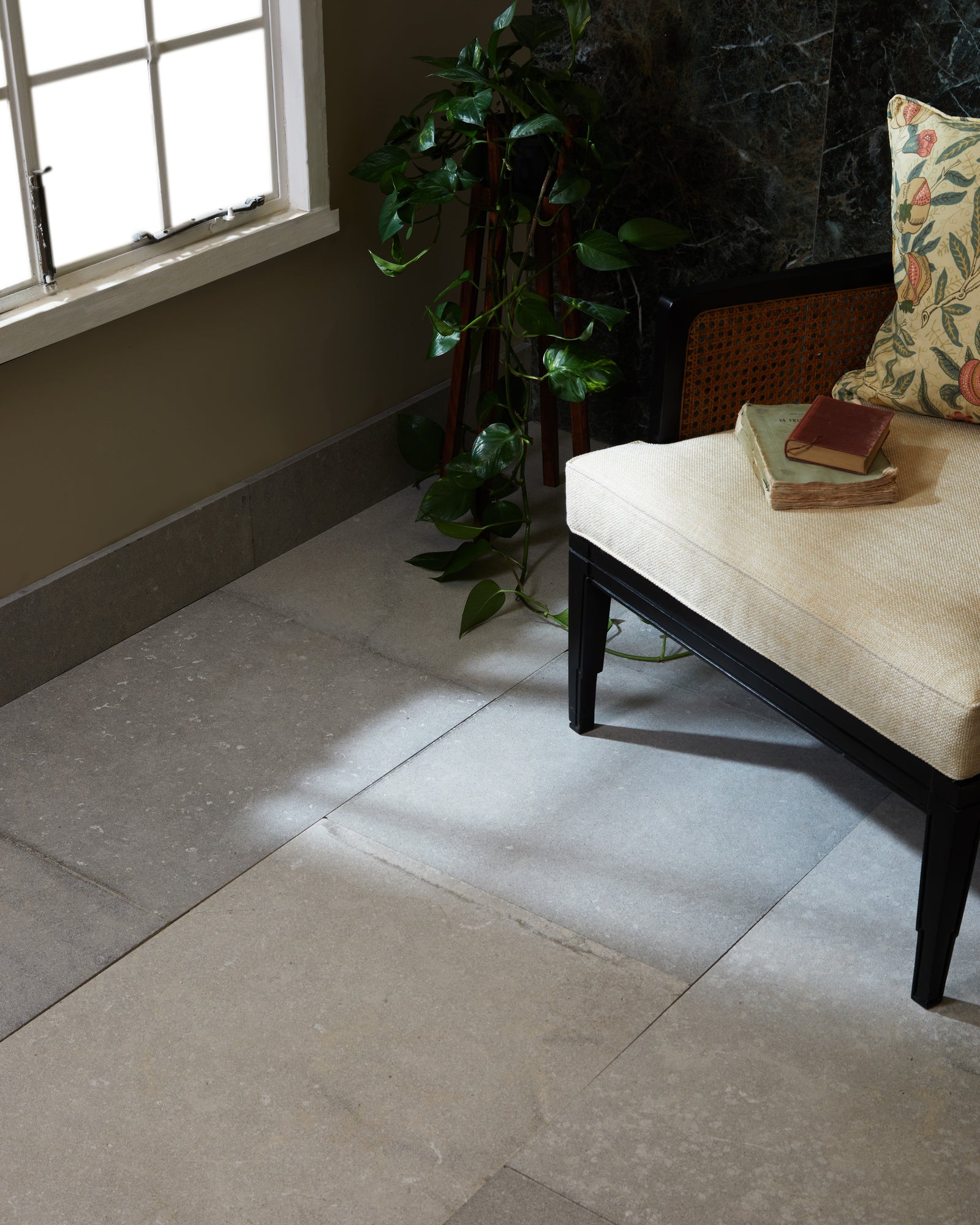Kimmeridge Limestone Etched Finish in large format underfoot, catching low sunlight beside a black cane bench, trailing pothos and a dark green marble wall