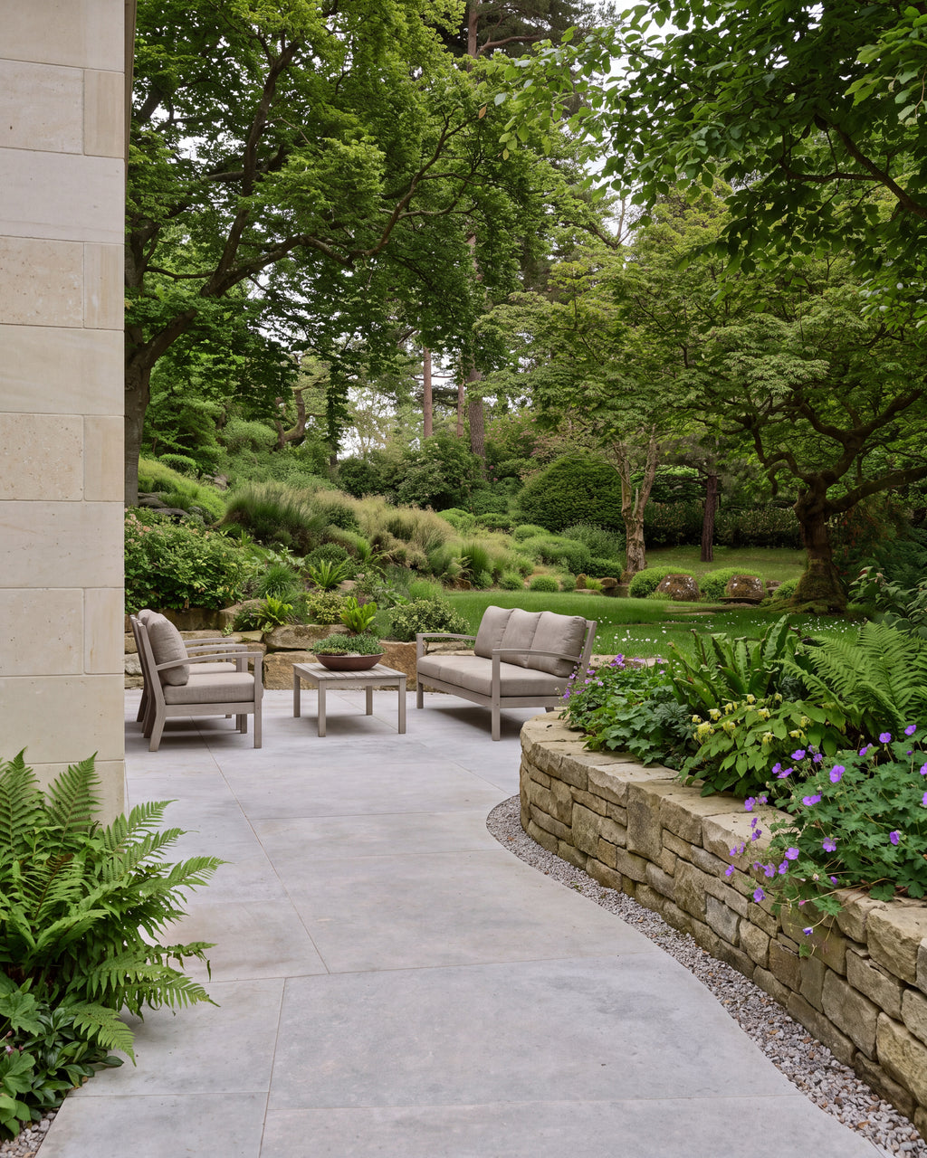 Kimmeridge Limestone Paving Etched Finish across a sheltered terrace with grey outdoor sofas, curving past a dry stone retaining wall planted with ferns and geraniums