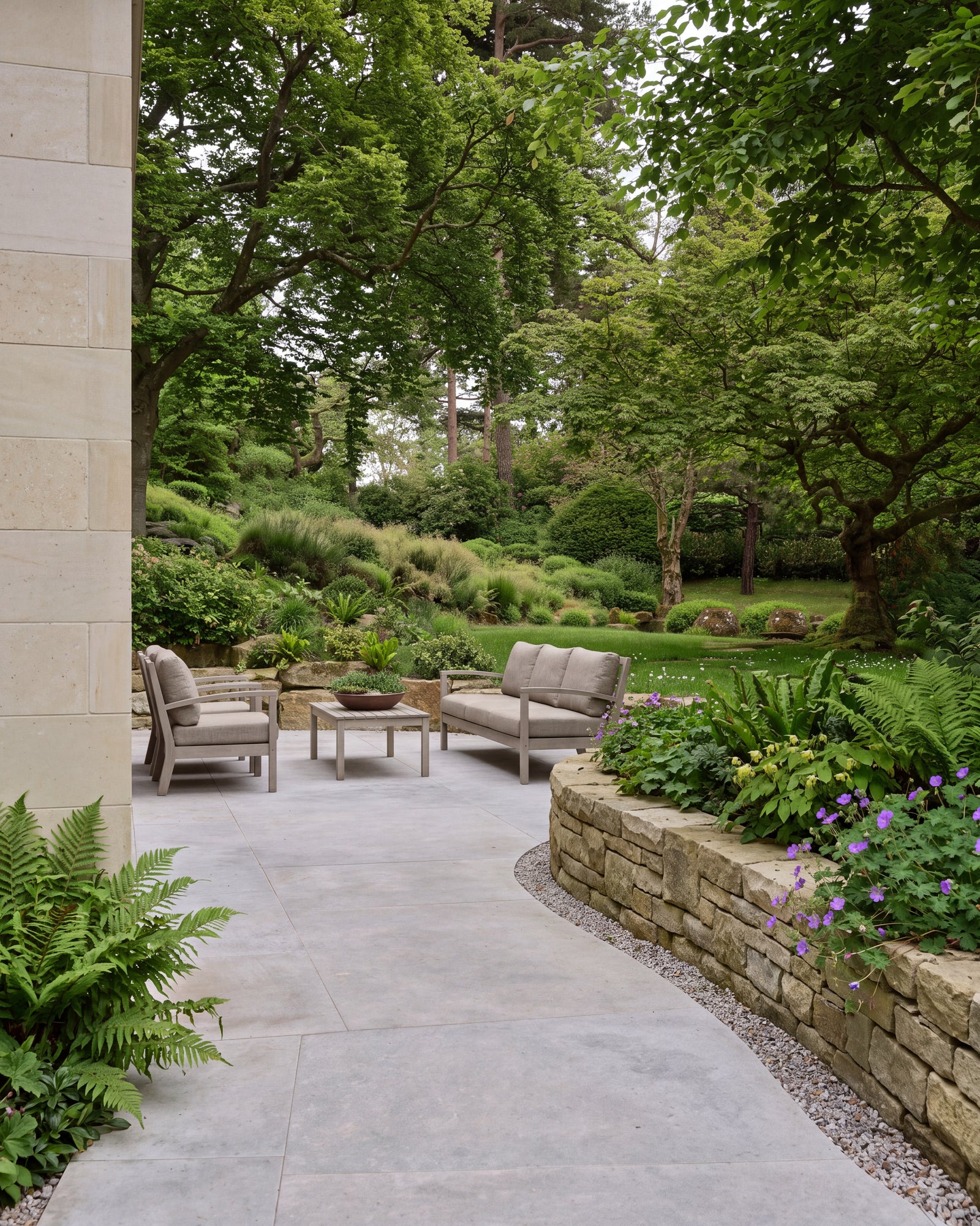 Kimmeridge Limestone Paving Etched Finish across a sheltered terrace with grey outdoor sofas, curving past a dry stone retaining wall planted with ferns and geraniums