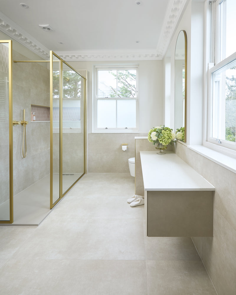 Loft Porcelain Pearl on the floor and walls of a long bathroom with a brass framed walk-in shower, floating vanity and arched mirror