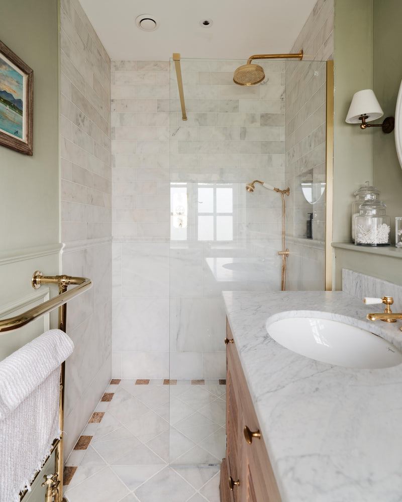 Long Island Marble Honed Finish on the floor and lower walls of a narrow sage green bathroom, with Ravenna Marble Honed Cabochon insets in the floor and a brass walk-in shower