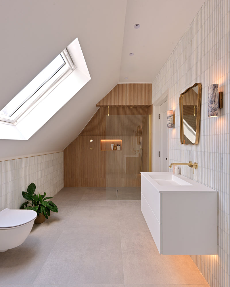 Louvre Porcelain Perla fluted tile running full height behind a floating white vanity and as a dado under the eaves, in a loft bathroom with a skylight, brass mirror and marble wall lights
