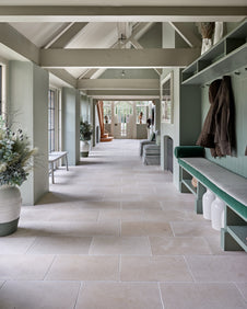 Mallory Limestone Tumbled Finish running the length of a green panelled boot room with a velvet bench seat and coat hooks