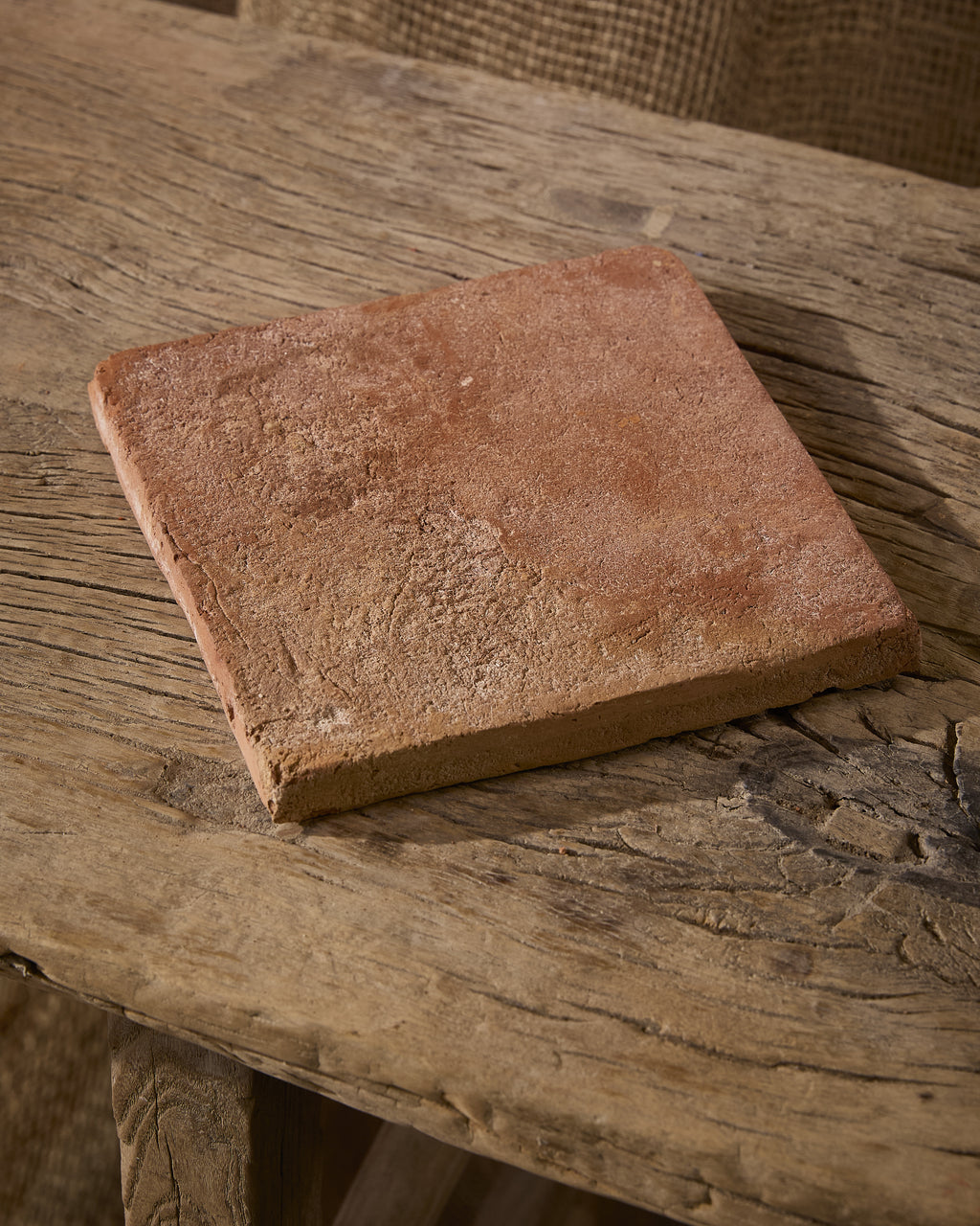 A single Marlborough Terracotta Square tile resting on a weathered elm bench, showing its warm terracotta face, chalky bloom and hand-formed edges
