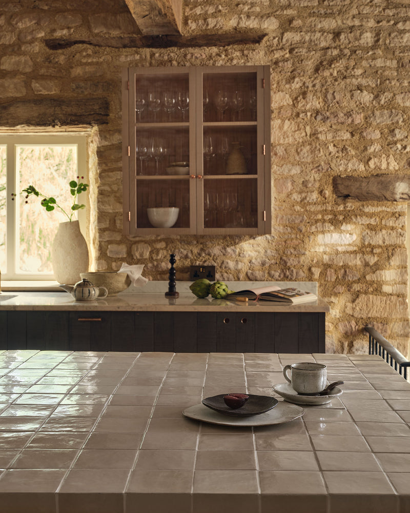 Maroc Ceramic Beige square glazed tiles clad across a kitchen island worktop in a rustic stone barn conversion, with an exposed beam and a glazed wall cabinet behind