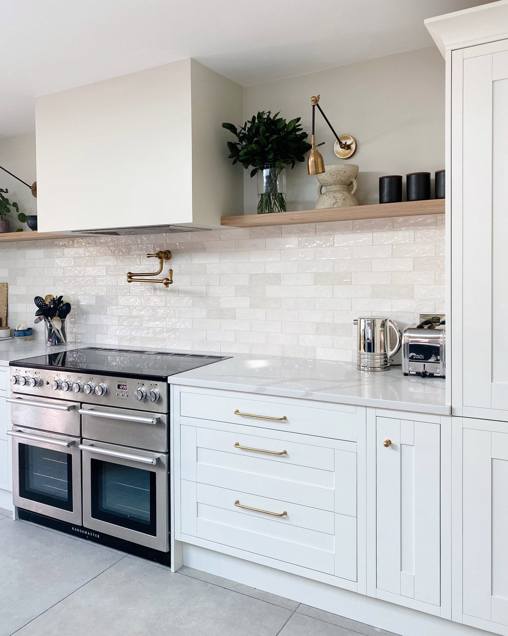 Oasis Ceramic White Gloss in a brick bond splashback running the length of a white shaker kitchen with brass handles, an oak shelf and a Rangemaster cooker